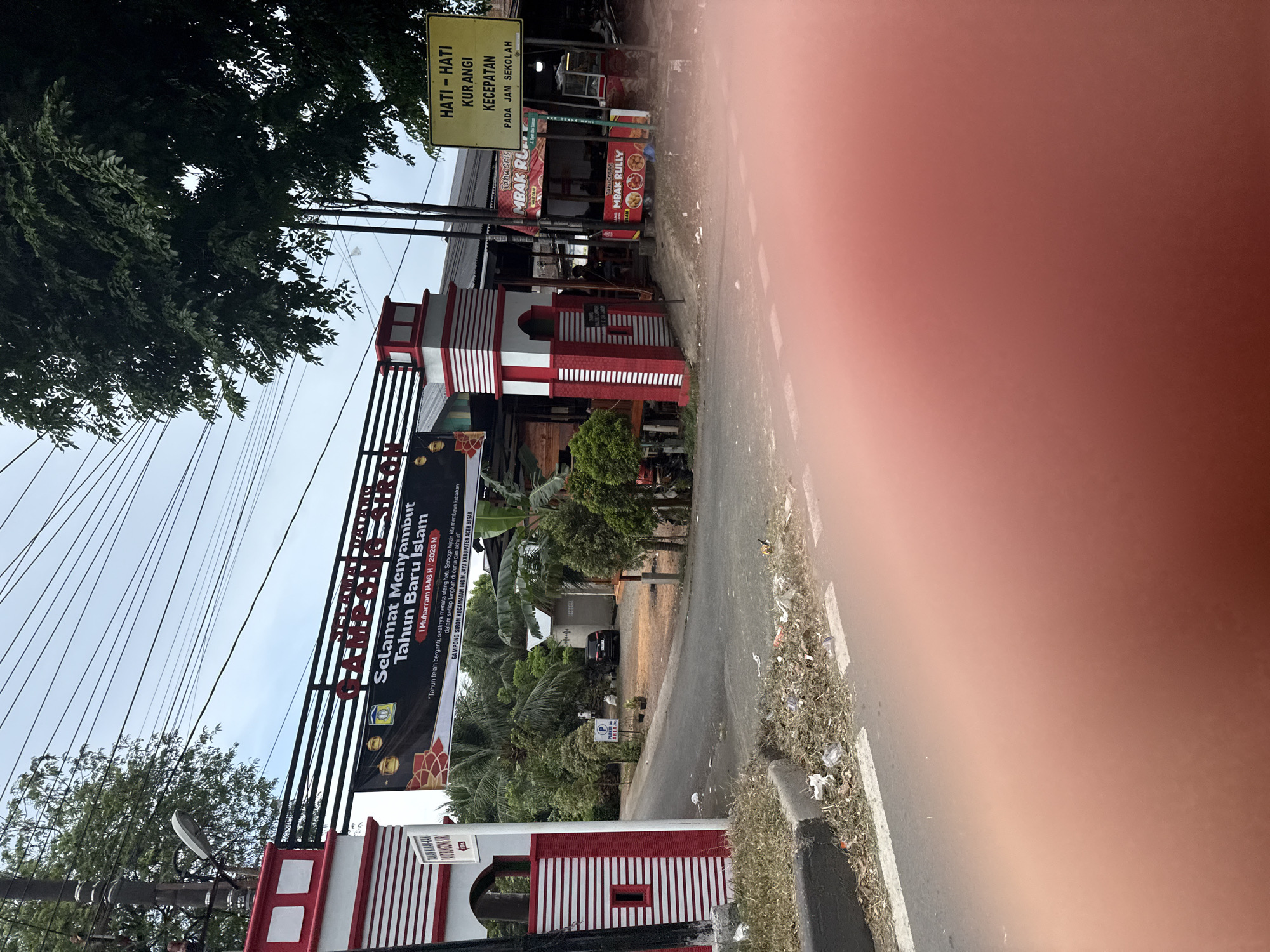 The image shows an outdoor scene in the city of Siron, Indonesia, with an arched gateway in the foreground. The gateway is red and white and has a banner that reads "SELAMAT DATANG GAMPONG SIREN" (Welcome to Gampong Siren) and "Selamat Menyambut Tahun Baru Islam" (Happy Islamic New Year). There are trees and buildings in the background. A yellow sign on the right reads "HATI-HATI KURANGI KECEPATAN PADA JAM SEKOLAH" (Be careful, reduce speed during school hours). Trash is visible on the side of the road. The sky is overcast.