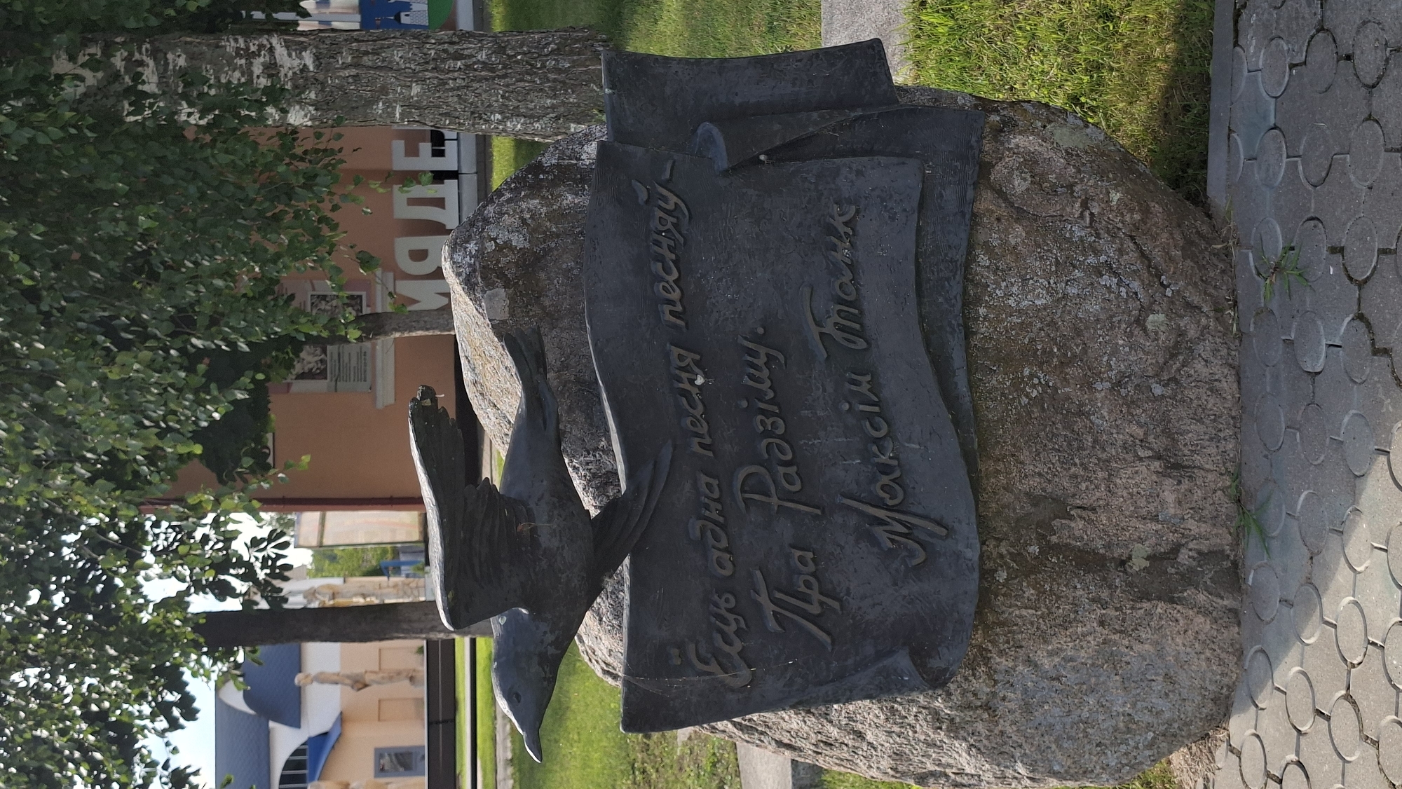 A weathered bronze sculpture of a bird in flight is mounted on a large boulder. The sculpture is positioned on a paved area with a grassy background. The boulder has an inscription in Cyrillic script that reads "Есть одна песня песняў, пра Радзіму. Максім Багдановіч" which translates to "There is one song of songs, about the Motherland. Maxim Bogdanovich." The sculpture is located in the city of Myadzyel, Belarus, and is likely a monument to the Belarusian poet Maxim Bogdanovich. The surrounding environment includes trees, a building in the background, and the distinct circular paving stones in the foreground. The overall scene suggests an outdoor public space dedicated to cultural heritage.