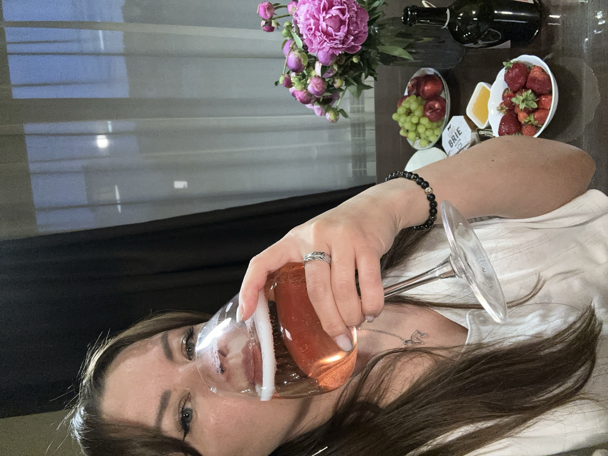 A woman with long, dark brown hair and winged eyeliner is holding a glass of pink sparkling wine to her lips. She is wearing a white shirt and a silver ring with a feather design on her right hand, and a black beaded bracelet on her left wrist. In the background, on a dark brown table, there is a vase of pink peonies, a bowl of green grapes and red plums, a small dish of honey, a piece of brie cheese, a bowl of strawberries, and a dark green bottle. The scene appears to be indoors, possibly a dining room, with sheer curtains visible through a window in the background, where the sky is visible. The location is the city of Chaikovsky, Russia.