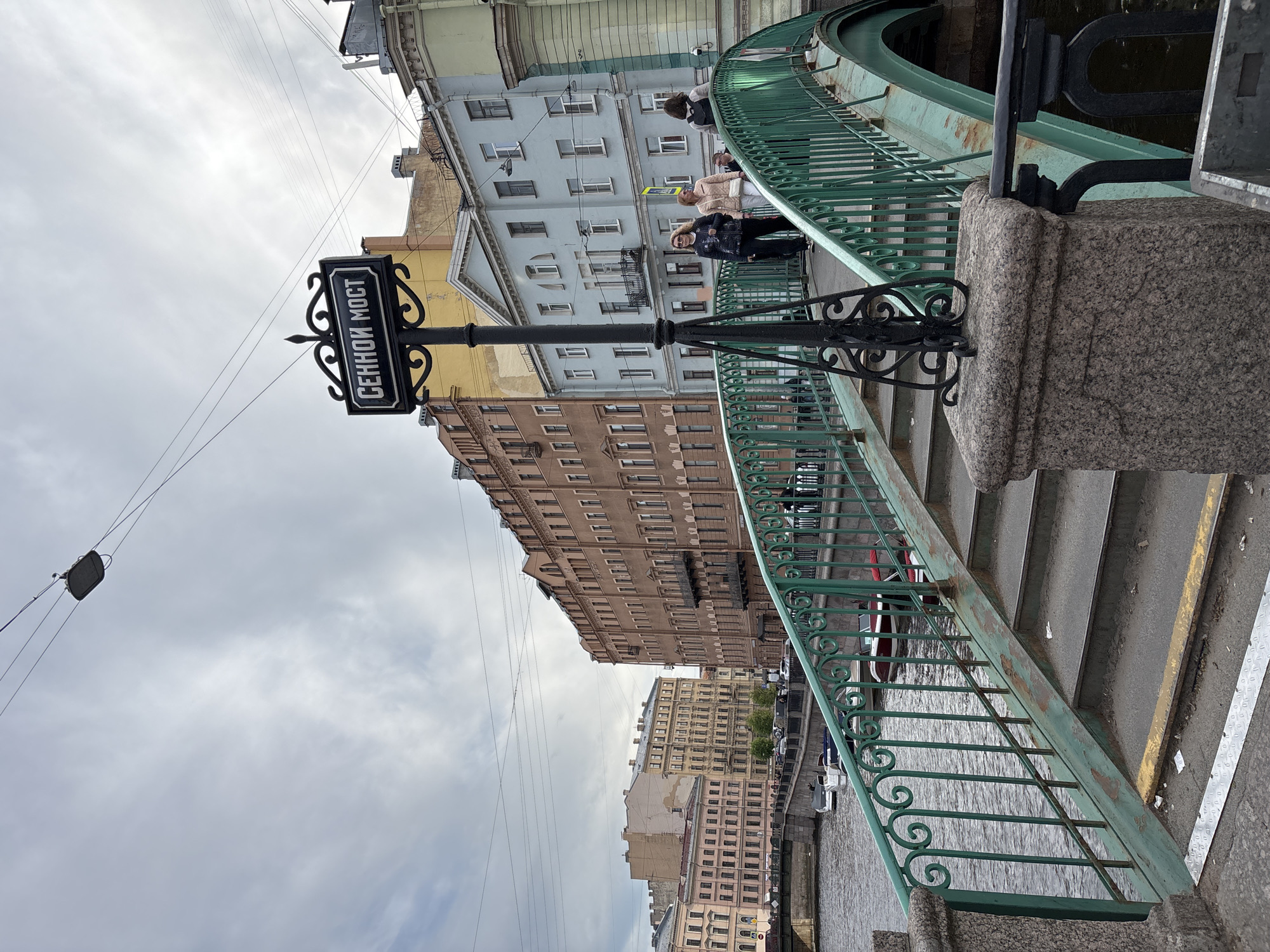 The image captures an urban landscape featuring a bridge over a canal or river, flanked by multi-story buildings, under an overcast sky. The photograph is taken from the bridge, looking along its length and towards the opposite bank.
In the immediate foreground, a sturdy green metal railing with decorative scrollwork patterns extends along the bridge's pedestrian walkway. The railing is anchored by robust, speckled grey granite pilasters. A prominent black sign with white Cyrillic text is mounted on an ornate black lamppost. The text clearly reads "СЕННОЙ МОСТ" (Sennoy Most), identifying the structure as Sennoy Bridge.
A few people are visible on the bridge's walkway. One person, dressed in a dark jacket, stands near the railing, while another is seen further down the bridge.
Beyond the bridge and its railing, the calm waters of the canal reflect the muted light of the cloudy sky and the facades of the surrounding buildings. A row of historic, multi-story buildings lines the canal banks, displaying a variety of architectural styles and colors, including light blue, yellow, and brown. These buildings feature numerous windows and some have small balconies.
The sky is entirely covered with a uniform layer of grey clouds, indicating a cloudy daytime setting. Utility wires crisscross the upper portion of the frame against the sky. On the right side of the image, concrete steps lead down from the bridge level, adjacent to a section of pavement marked with a yellow line. The scene depicts a typical day in an older European city, specifically Saint Petersburg, Russia.