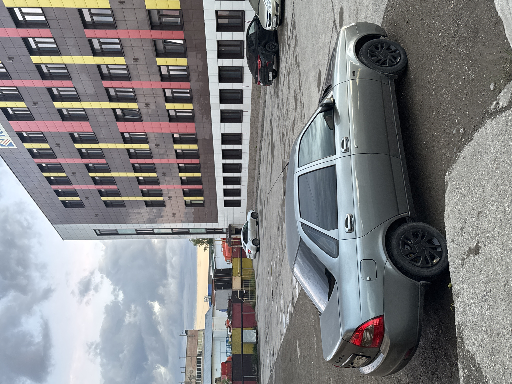 The image captures an urban street scene in Perm, Russia, under an overcast sky, suggesting recent rain given the wet asphalt. The perspective is tilted, showing a multi-story building to the left and parked cars to the right.
The main subject is a light grey sedan, likely a Lada, parked on the wet asphalt in the foreground. It features black multi-spoke wheels, and its rear license plate "Р569СА 159" is partially visible, indicating the Perm Krai region. Further back, another black sedan with license plate "У897РХ 159" is parked, and a white hatchback is visible beyond it. A fourth, silver/grey car is partially visible behind the black one.
To the left, a large, modern building dominates the background. Its facade is a mosaic of dark grey panels, accented by prominent horizontal stripes of bright yellow and red. The windows are dark-framed. A logo or text, possibly "ЛГД" in blue and white, is visible on the lower left section of the building. In the far background, behind the cars and to the left, industrial-type structures and stacked shipping containers (red, yellow, black) are visible, hinting at an industrial zone or storage area.
No people are visible, and there is no discernible activity taking place; the cars are all parked. The overall impression is a static, contemporary urban environment on a cloudy day.