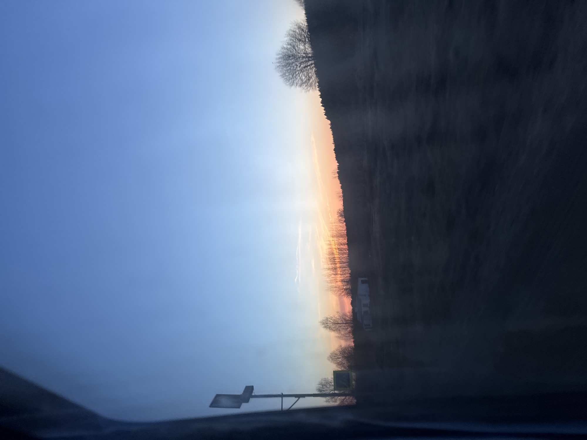 The image shows a view from inside a vehicle, likely a car, during sunrise or sunset. The sky transitions from a light blue to an orange-yellow horizon. In the distance, a treeline and what appears to be a building (possibly residential or commercial) are visible. There is also a traffic sign on the left side of the road. The perspective suggests the photo was taken while the vehicle was in motion. The location in Krasnye Gorki, Russia is not definitively ascertainable based on the visual information alone.