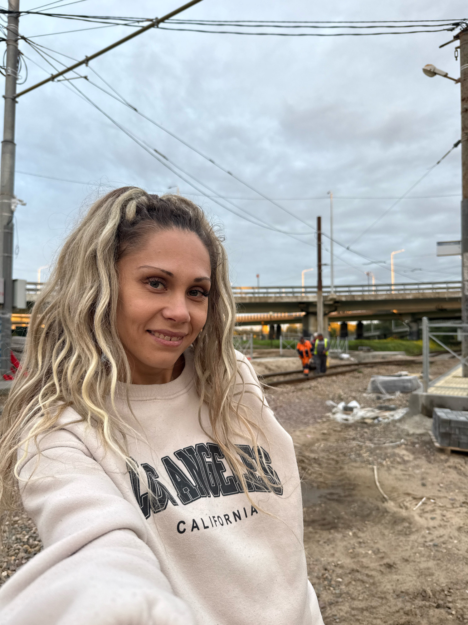 A woman with long, wavy blonde hair and light skin, wearing a cream-colored sweatshirt with "LA ANGELES CALIFORNIA" printed on it, poses for a selfie. She is smiling, with her head slightly tilted to the right. Her makeup is visible, including mascara and eyeliner. In the background, there is a construction site with railway tracks, gravel, piles of bricks, and building materials. Two men in orange and yellow safety vests are working on the tracks in the distance. Overhead, power lines and streetlights are present, and a bridge crosses in the background. The sky is overcast. The location is identified as Yaroslavl, Russia, likely a construction area near a transportation hub.