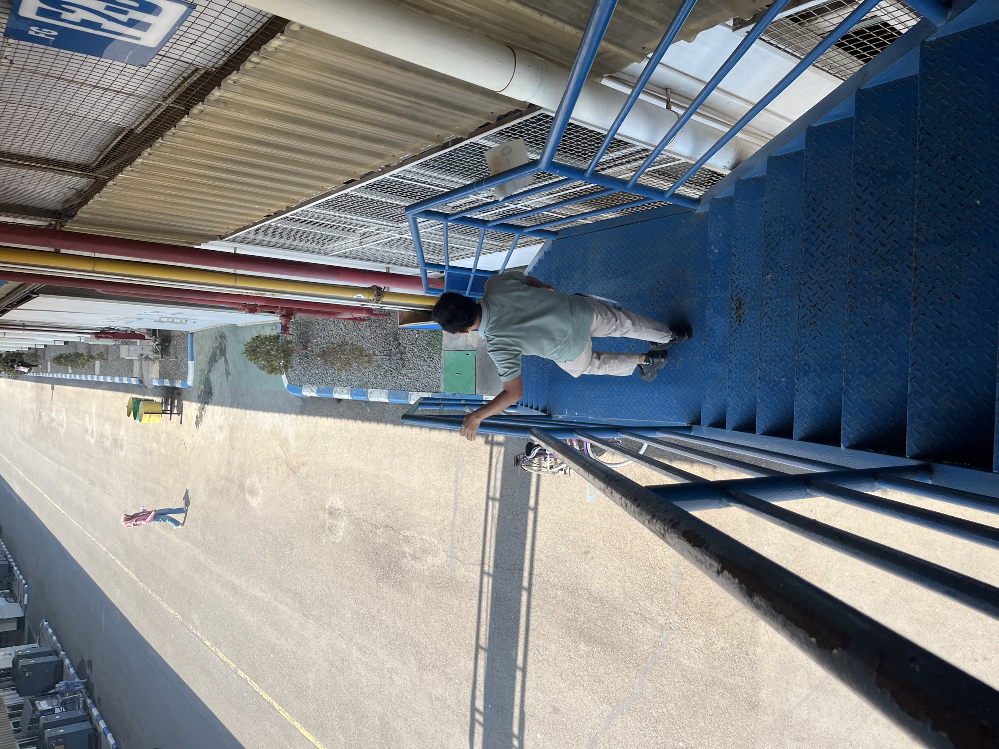 An overhead shot captures a man in a green polo shirt and tan pants descending a bright blue metal staircase, his right hand gripping the railing. He is walking down the stairs, facing away from the camera. The staircase has a diamond plate pattern on the steps and is bordered by a blue metal railing. The scene is bathed in bright sunlight, casting sharp shadows. To the left of the stairs, a paved area is visible, with a bicycle parked near the bottom of the frame. Further in the background, a woman wearing a pink hijab and blue jeans walks away from the camera on a wide paved pathway. The pathway is marked with a yellow line and has two green trash receptacles on its right side. Beyond the pathway are buildings with industrial features, including corrugated metal walls and various colored pipes. The overall environment suggests an industrial or port area, possibly a dock or factory. The location is identified as Pelang, Indonesia.