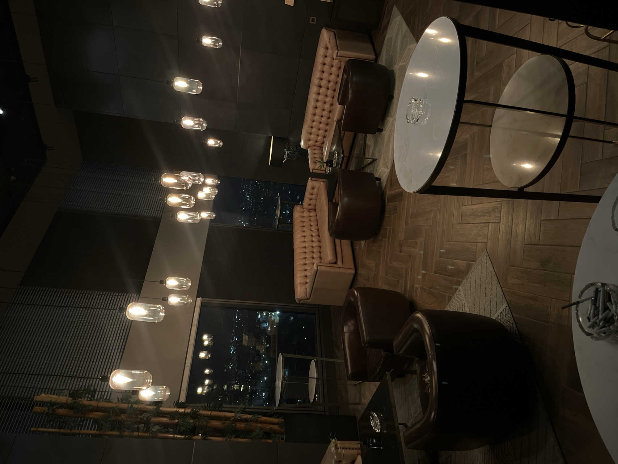 The media file presents an interior view of a sophisticated, dimly lit bar or lounge at night, likely situated in a high-rise building in Sofia, Bulgaria. The establishment offers a panoramic view of the city's illuminated skyline.
The scene is furnished with several seating arrangements, including tufted, light brown upholstered sofas or banquettes and dark brown leather armchairs. These are complemented by various round tables featuring light-colored tops and dark metal bases; some are high-top tables, while others serve as lower coffee tables. An ashtray is visible on one of the tables.
Numerous exposed filament pendant lights hang from the ceiling, emitting a warm, inviting glow that illuminates the space. The floor is composed of light-toned wooden planks arranged in a geometric pattern, with a patterned rug partially covering the area under some seating. Large windows frame the view of a bustling city at night, with countless lights from buildings and streets. A decorative element resembling bamboo stalks with some greenery is positioned near one of the windows.
No people are visible in the image, suggesting the scene is quiet or captured before or after peak hours. There is no specific activity taking place. The time of day is clearly night, as indicated by the dark sky outside and the reliance on interior lighting. No legible text is visible in the image.