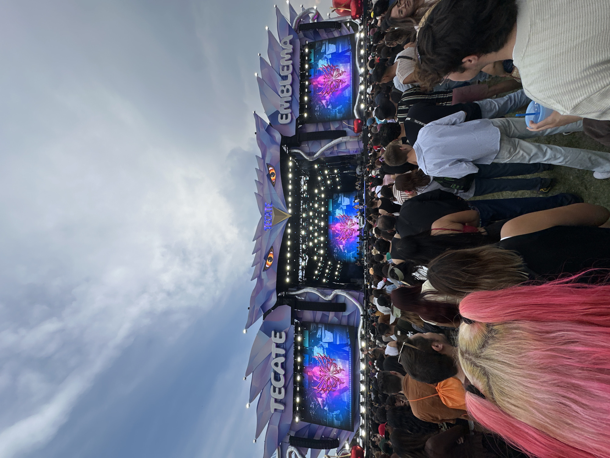 The image captures a crowded outdoor event in Mexico City, Mexico. A large stage with the words "EMBLEMA" and "TECATE" is visible, displaying a floral design on its screens. The audience, composed of numerous individuals, faces the stage. Some people are sitting on the grass, while others are standing. A few have brightly colored hair. The sky above is partially cloudy.