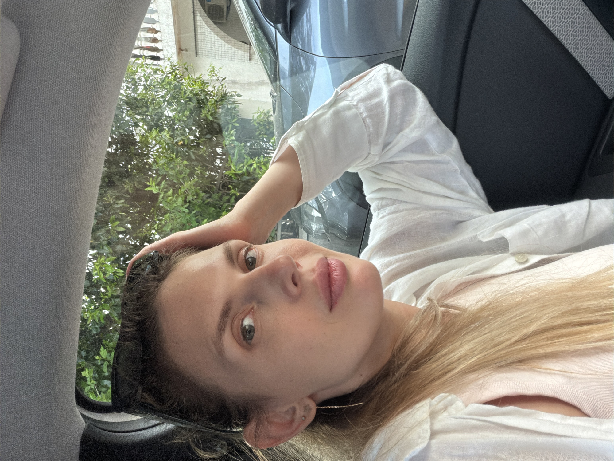 A woman is lying in a car, possibly a taxi, with her head on her hand. She is wearing a white linen shirt over a pink top. Her blonde hair is visible near her chest. The car seat is black leather and the seatbelt is gray. Outside the car window, there is greenery, likely a tree or bushes, and a glimpse of a building in the city of Budva, Montenegro.