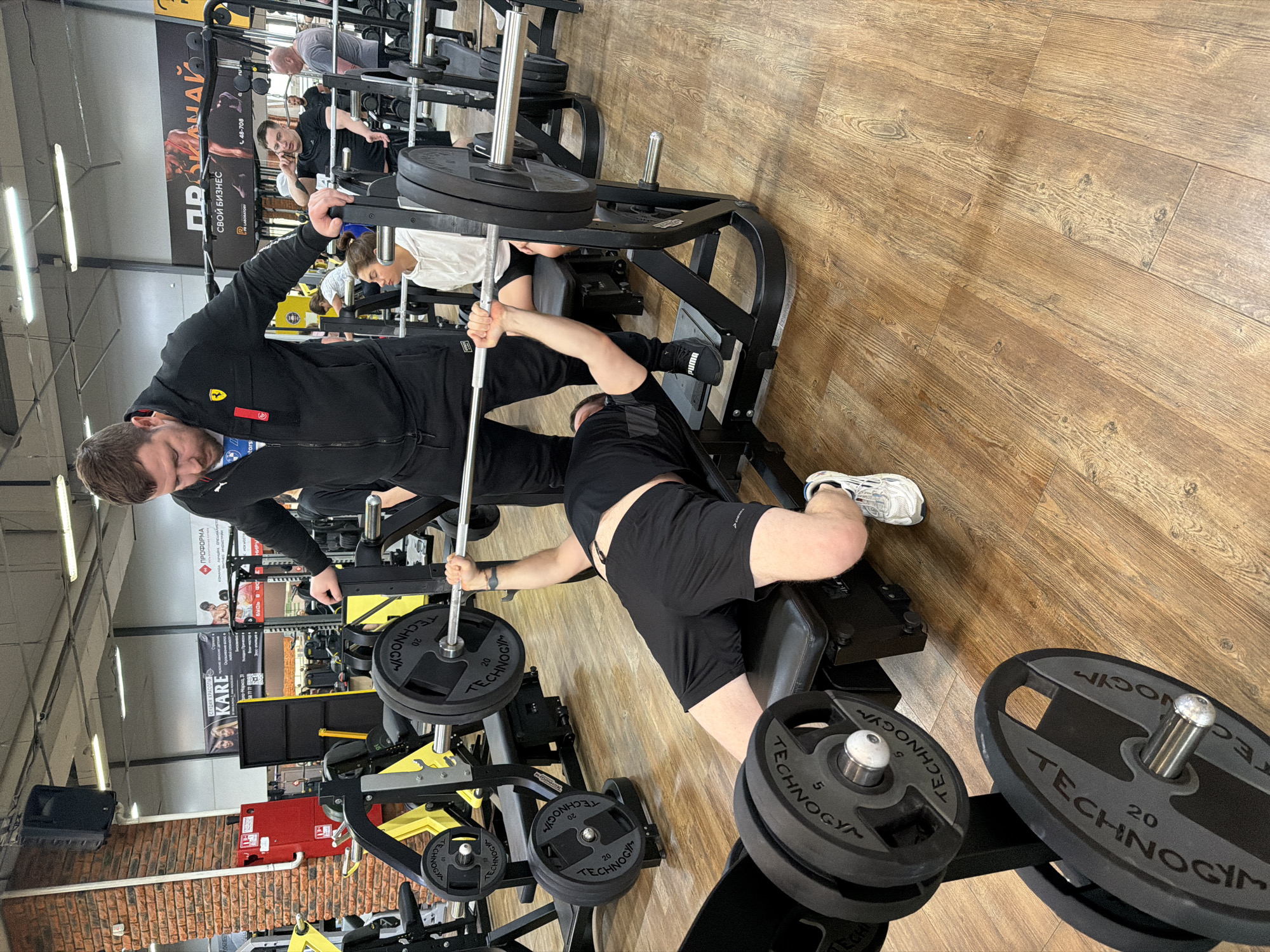 The scene is an indoor gym or fitness center, located in Unknown, Unknown. In the foreground, a male individual is performing a supine barbell bench press on a black padded bench. He wears a black t-shirt, black shorts, and white athletic shoes, with his feet on the wooden-patterned floor. A second male, identified as a spotter, stands over him, wearing a black Ferrari-branded zippered hoodie, black athletic pants, and black Puma shoes; his hands rest on the upright supports of the black and yellow bench press machine. The barbell is loaded with several Technogym weight plates, including visible 20kg and 5kg discs. In the mid-ground, a female in a white hoodie sits on a bench, while another male in a black t-shirt stands, holding a mobile phone to his ear. The background reveals additional gym equipment, mirrors, and wall-mounted banners featuring Cyrillic text, specifically "СТУДИЯ КРАСОТЫ KARE" (Beauty Studio KARE) and "СВОЙ БИЗНЕС" (YOUR BUSINESS). A red fire safety cabinet is affixed to a prominent brick column. Overhead, industrial-style lighting illuminates the facility.
