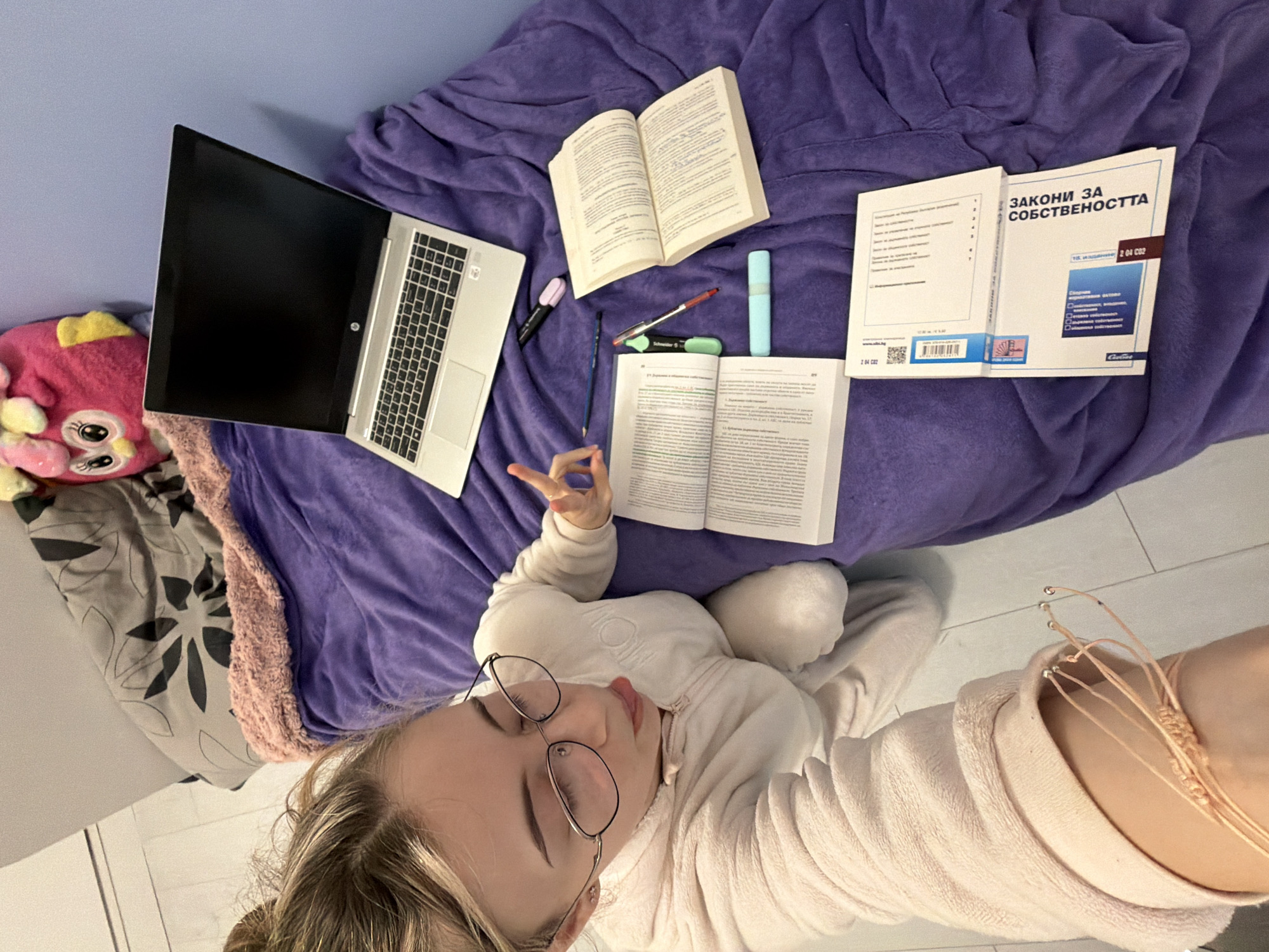 A high-angle selfie shows a person lying on a purple blanket on a bed or couch, surrounded by study materials. A partially open laptop is to the left, and three books, pens, and a highlighter are scattered around. One book is open, and another is titled "ЗАКОНИ ЗА СОБСТВЕНОСТТА" (Laws of Ownership), suggesting the person is studying law. A plush toy is visible in the lower left corner. The person is wearing glasses and a light-colored, fuzzy garment. Based on the book title, this could be taking place in Bulgaria. However, without further evidence or information, determining the exact location, including the city of Plovdiv, Bulgaria, is not possible.