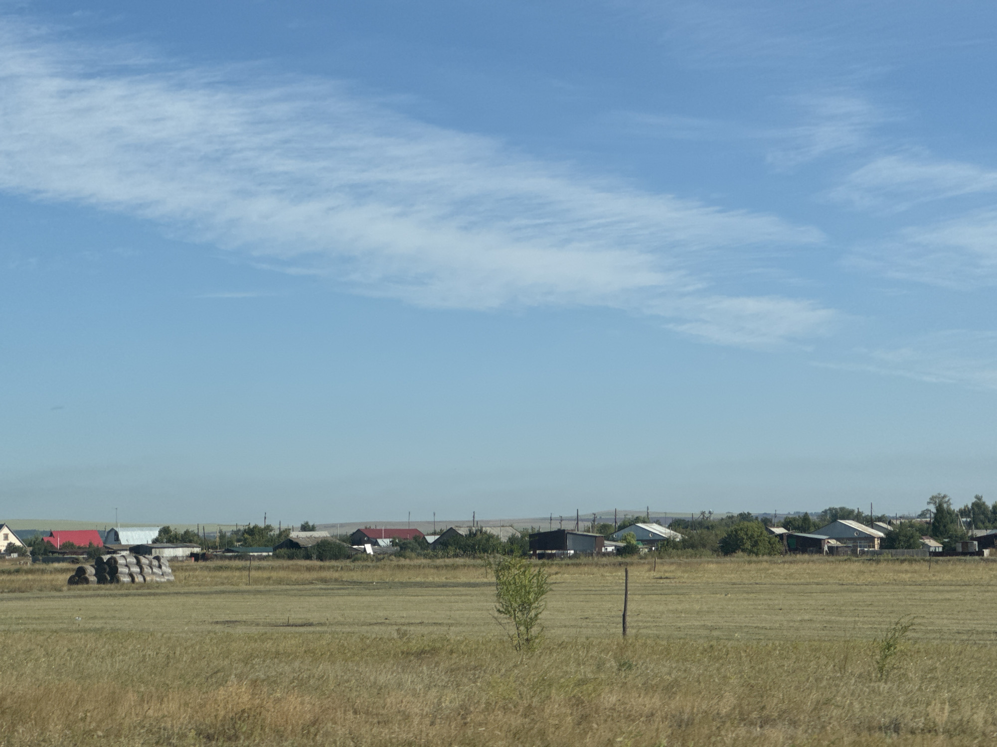 In the city of Gamaleevka-1, Russia, an exterior shot shows a rural landscape under a clear blue sky with scattered cirrus clouds. In the foreground, a field of dry, golden grass stretches across the image. In the middle ground, a cluster of hay bales is visible to the left, and a small, green tree stands in the center, with a wooden post to its right. A line of buildings, appearing to be houses and sheds, forms a village in the background. The buildings have various roof colors, including red, white, and dark gray. The overall scene is one of a quiet, rural settlement on a bright day.