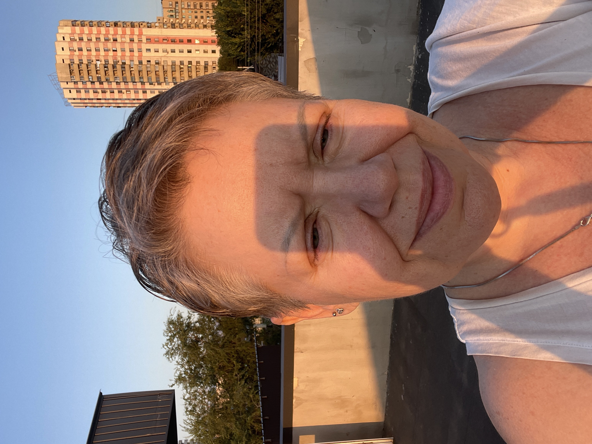 A middle-aged woman with short, graying hair is taking a selfie outdoors at sunset. She is wearing a white tank top and a silver necklace. The light of the setting sun casts a warm glow on her face and body, creating a strong shadow across her forehead and nose. In the background, there is a building with a distinctive pink and beige facade, and other buildings and trees are visible. The woman's facial expression is relaxed, with a slight smile. The scene appears to be on a rooftop or elevated area, with a concrete wall and a dark surface in the foreground. The overall impression is a candid moment captured during golden hour. The architectural style of the buildings suggests an urban environment, possibly in Almaty, Kazakhstan, given the presence of the distinctive residential tower.