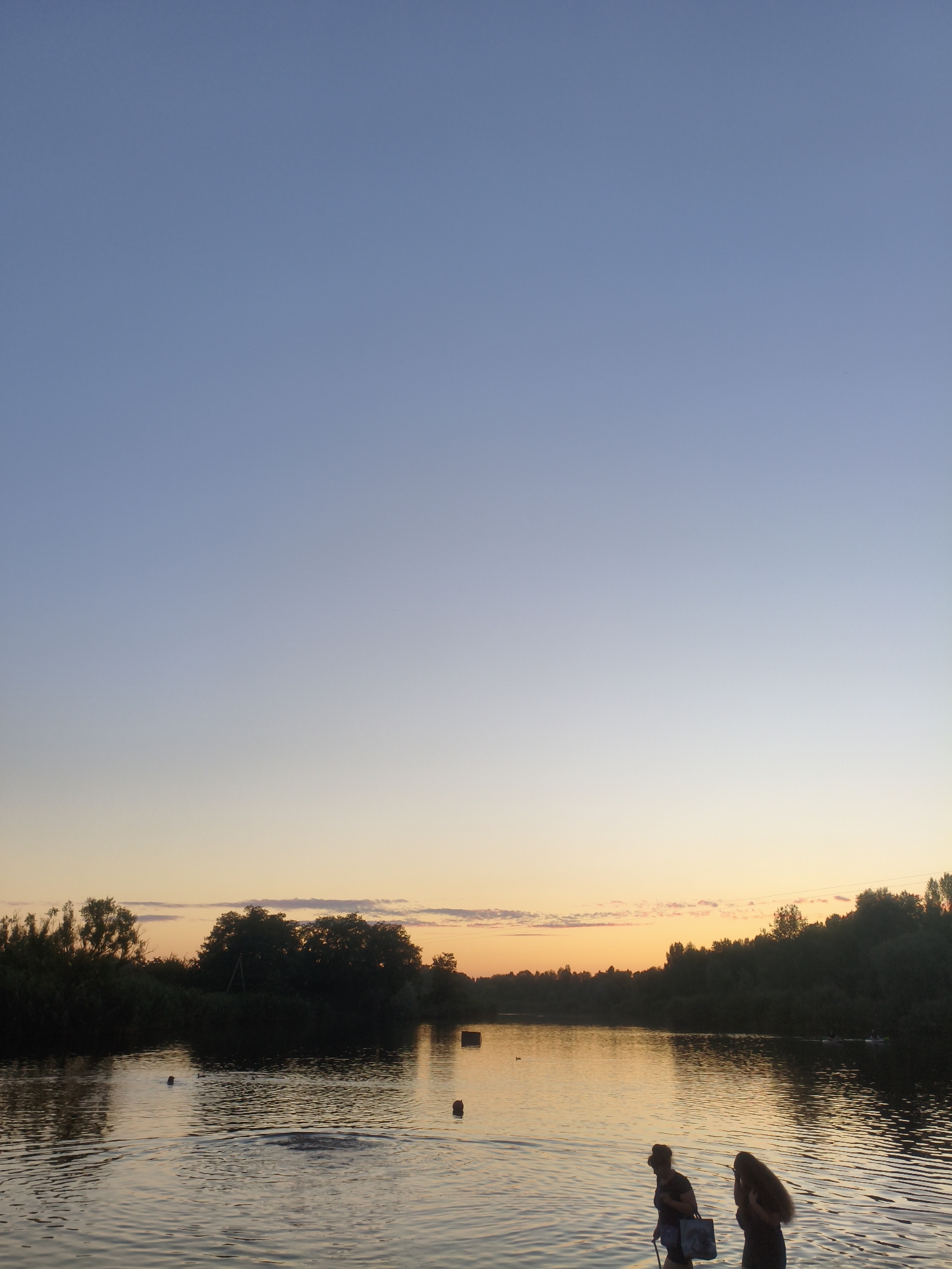 At sunset on a body of water, possibly a river or lake, two individuals, one wearing a hat and carrying a bag, and another with long dark hair, walk along the shore. The sky transitions from a clear blue at the top to a soft orange and yellow horizon. The water reflects the sky and surrounding trees, which form a dark silhouette against the lighter sky. Several buoys are visible in the water, and there are faint ripples indicating movement. The scene suggests a peaceful evening by the water, with no specific urban landmarks identifiable to pinpoint a location within Kremenchuk, Ukraine.