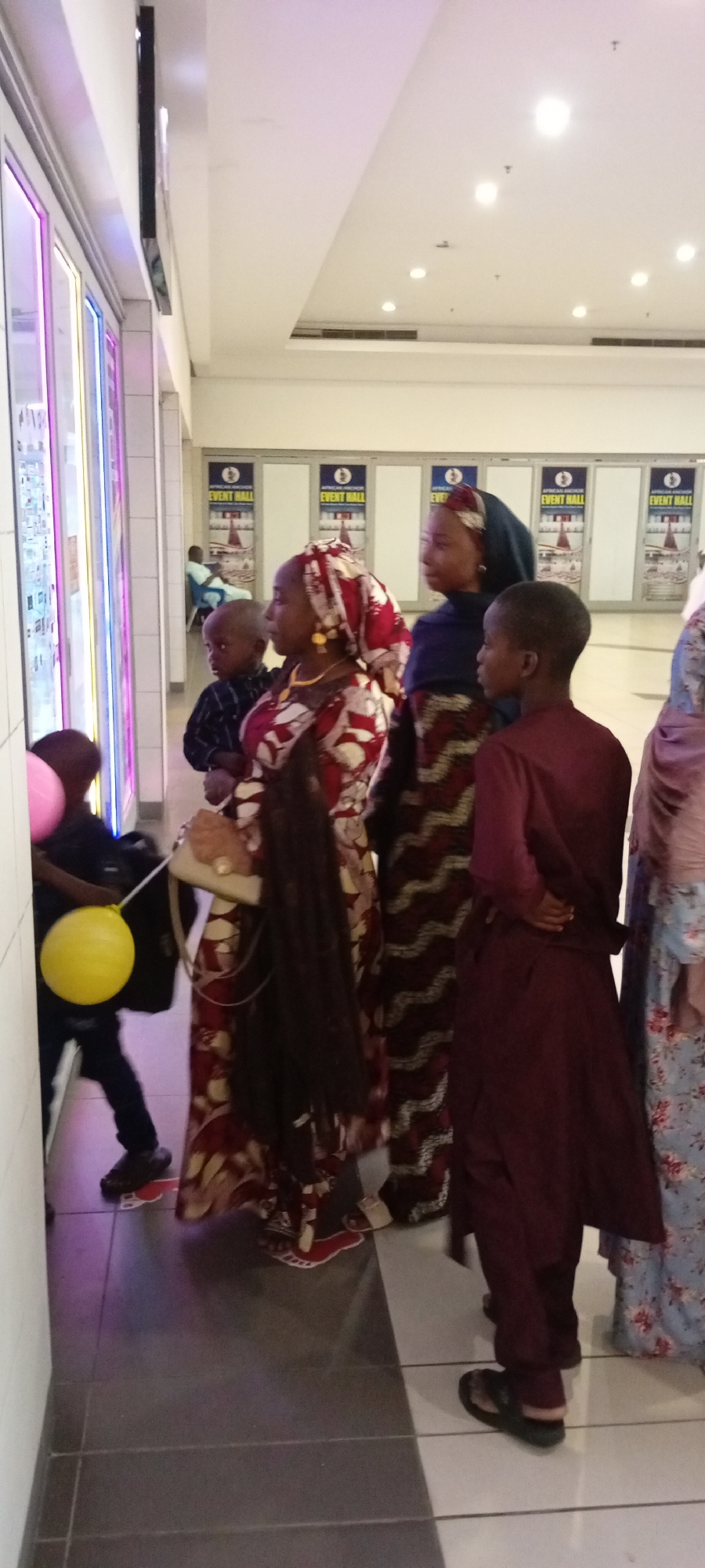 In Gandu Albasa, Nigeria, a group of individuals, including children and adults, are gathered in an indoor environment, possibly a shopping mall or event center. The scene is characterized by bright lighting from the ceiling and the illuminated displays along a wall on the left. Several people are dressed in traditional African attire, featuring vibrant patterns and headscarves.
In the foreground, a young boy in dark clothing holds a yellow balloon tied to his wrist, while a pink balloon is also visible nearby. To his right, a woman in a patterned red and white dress and headscarf is looking towards the displays. Another woman, wearing a blue headscarf and a patterned dress, stands beside her. Further to the right, a child in a maroon outfit stands facing away from the camera. A woman in a floral-patterned dress is partially visible at the far right.
In the background, several doorways are visible, each marked with "AFRICAN ANCHOR EVENT HALL" and a circular logo. A person is seated in one of these doorways. The floor is tiled with a dark and light grey pattern, with red and white foot-shaped markings on the floor, possibly indicating social distancing guidance. The environment appears to be a public indoor space with a clean and well-maintained appearance.