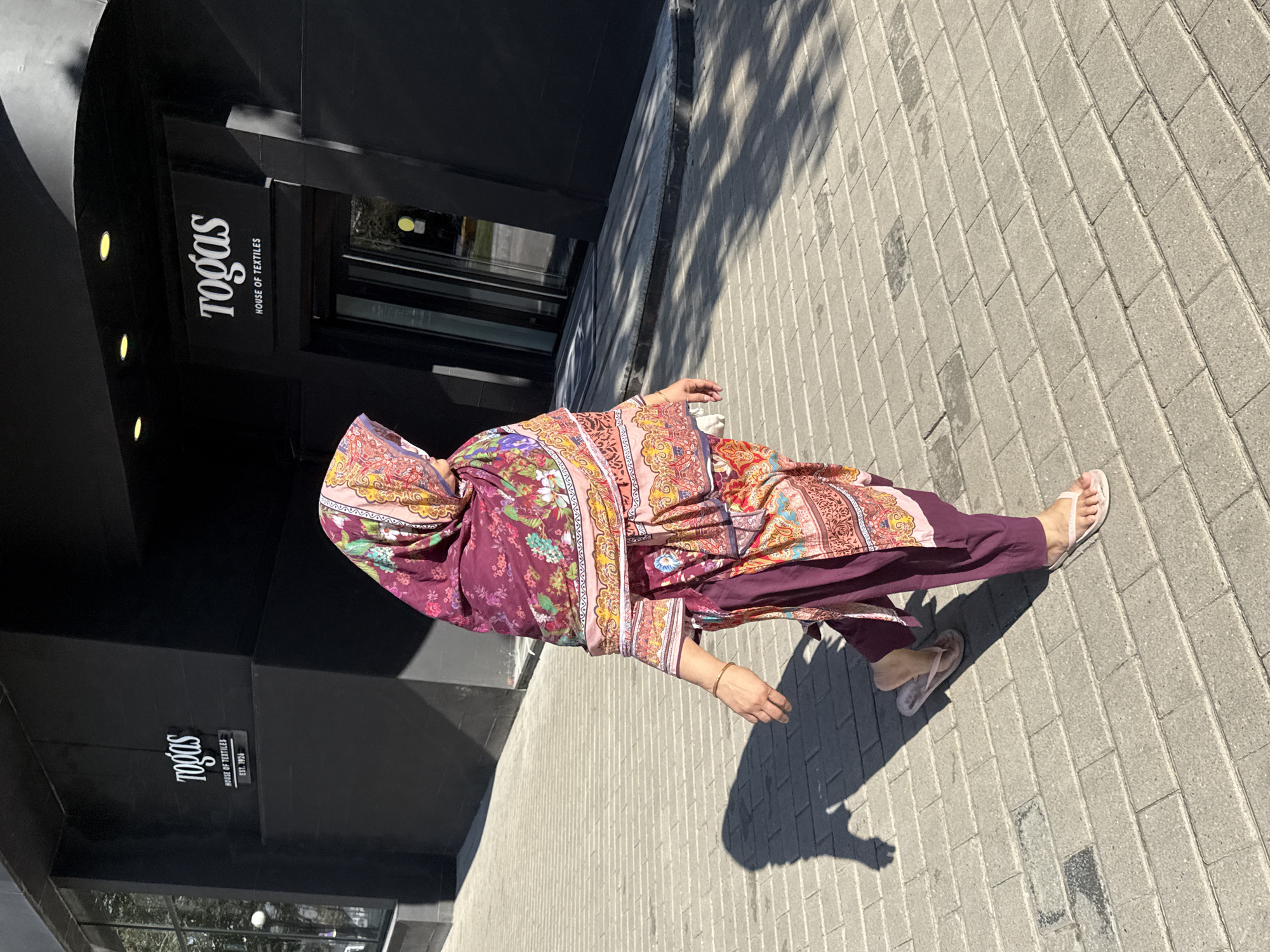 A woman wearing a vibrant, patterned headscarf and a matching outfit walks across a paved walkway in front of a modern, black building. The building has signs that read "togas" and "HOUSE OF TEXTILES EST. 1926". The walkway is made of grey rectangular pavers. The woman is wearing light pink flip-flops. Her shadow is cast on the walkway to her left. The sun is shining, creating dappled shadows from the building onto the walkway. The scene appears to be in a city environment, possibly a commercial district. The overall impression is one of a person walking on a sunny day in front of a retail establishment.