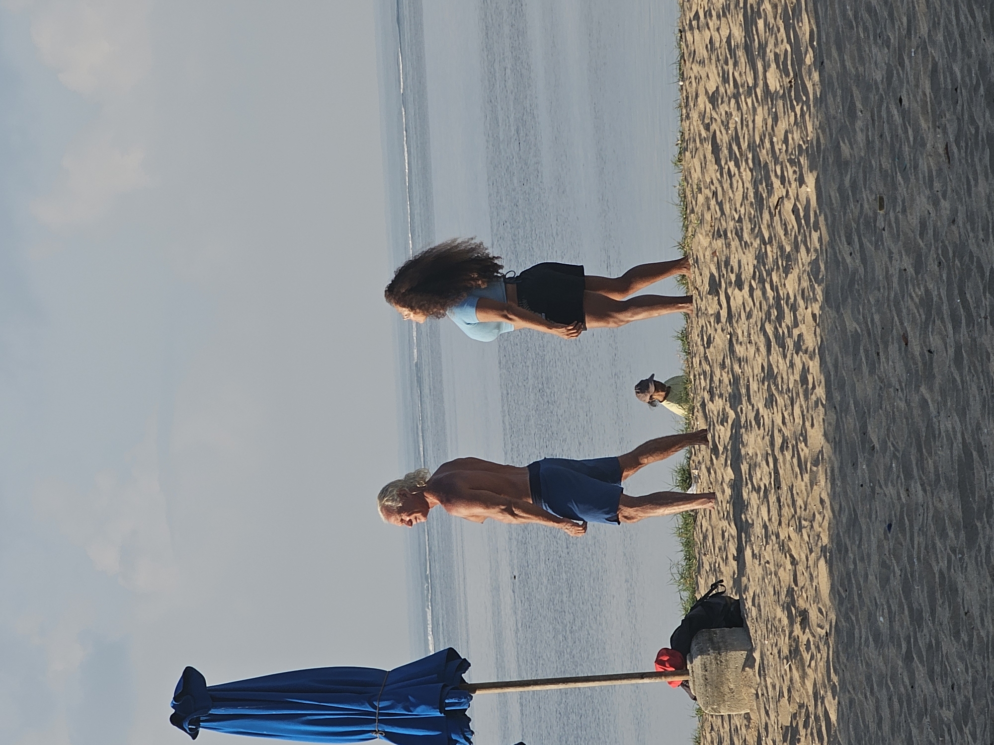 At Kuta Beach in Denpasar, Indonesia, a muscular man with blonde hair tied back in a ponytail and a muscular build stands on the sandy shore, wearing blue swim trunks. He is facing away from the camera, looking out at the ocean. Beside him, a woman with long, dark curly hair is also looking out at the ocean. She is wearing a blue crop top and black shorts. In the background, the ocean stretches to the horizon, with gentle waves. A closed blue beach umbrella is planted in the sand to the left of the man. A small child wearing a hat is seated on the sand in the middle ground, to the right of the man. The beach is covered in light-colored sand with some sparse green grass growing near the water's edge. The sky is a pale blue with some wispy clouds.