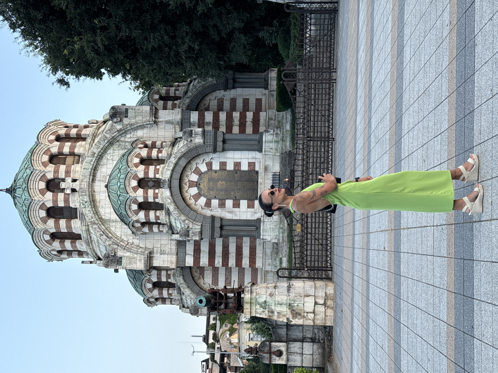 The image depicts a woman in a lime green dress standing on a tiled plaza in front of the St. Nicholas Chapel Mausoleum in Pleven, Bulgaria. The mausoleum is a prominent white and red striped domed structure with multiple arched windows. To the left of the mausoleum, there is a stone pedestal with an old cannon. The woman is facing right, looking away from the camera, with sunglasses on and her hair pulled back. She is wearing white wedge sandals and carrying a black purse. The background includes trees and a clear sky.