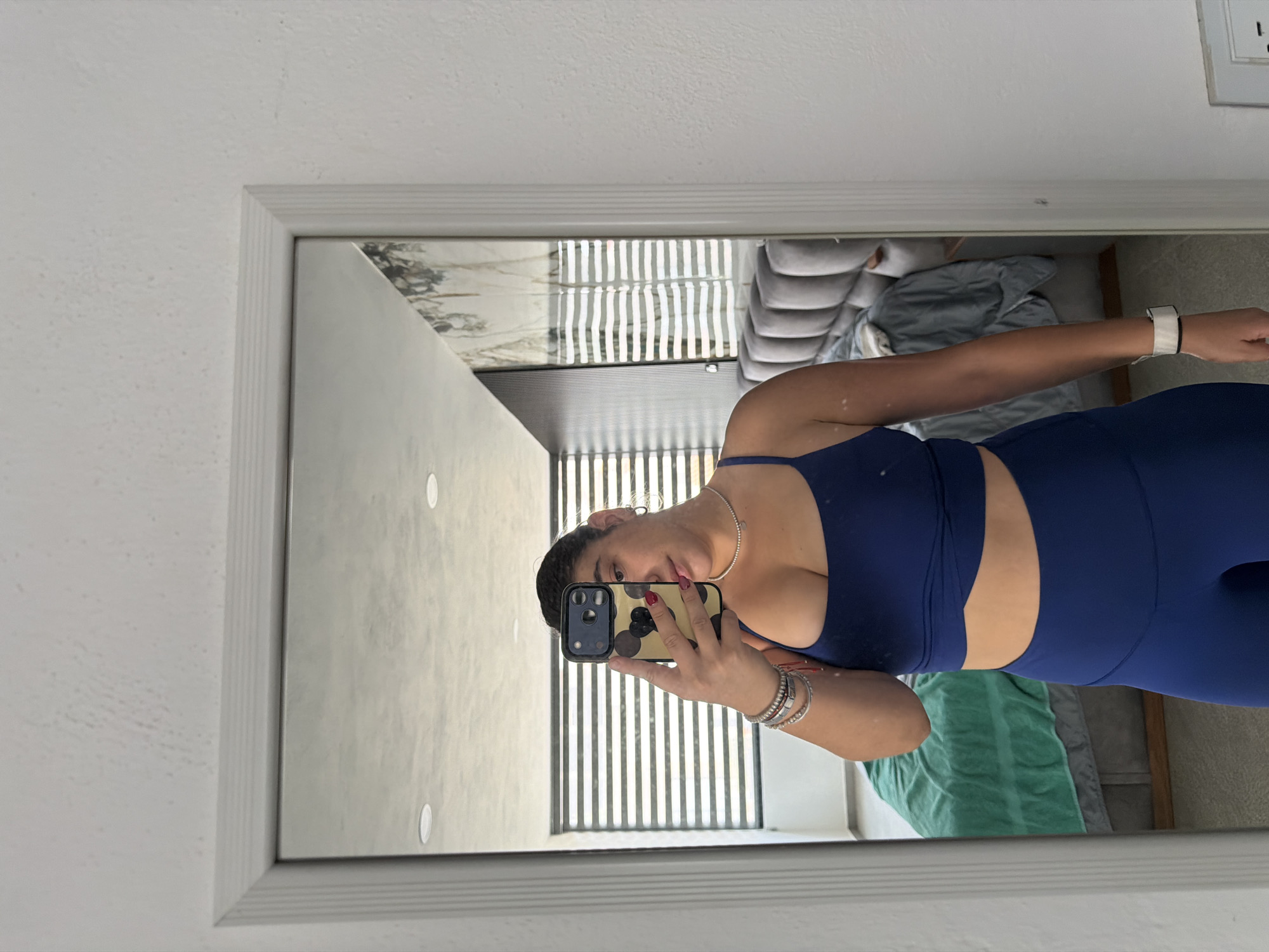 A woman is taking a selfie in front of a mirror. She is wearing a blue workout set and has several bracelets on her left wrist. Her iPhone has a patterned case. The mirror reflects a bedroom with a bed, curtains, and what appears to be an outlet on the wall. The location is Puebla, Mexico, based on the metadata (not visible in this specific cropped image) that indicates the photo's origin.