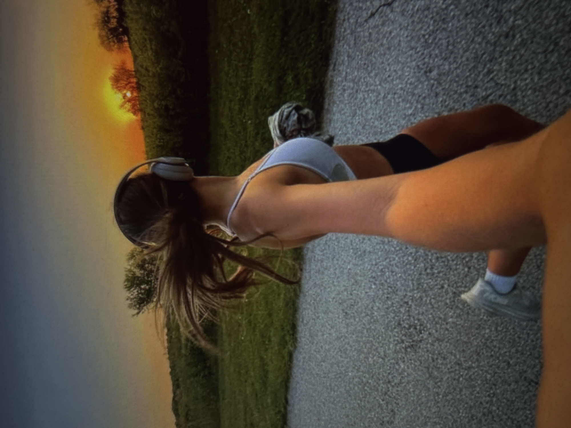 The media file shows a person from the chest down, wearing a white sports bra, black athletic shorts, white socks, and grey athletic shoes. They are wearing over-ear headphones and have long, dark hair tied back in a ponytail. The person is captured mid-stride on a grey asphalt path, suggesting they are jogging or running.
The background depicts a grassy field bordering a wooded area with trees. The sun is low in the sky, casting a warm orange and yellow glow, indicating either sunrise or sunset. The lighting suggests pleasant weather conditions. The scene is set in a natural, outdoor environment, possibly a park or a trail, given the grassy area and trees.
There is no visible text in the media file. The overall impression is one of an active individual enjoying an outdoor workout during a beautiful time of day.
