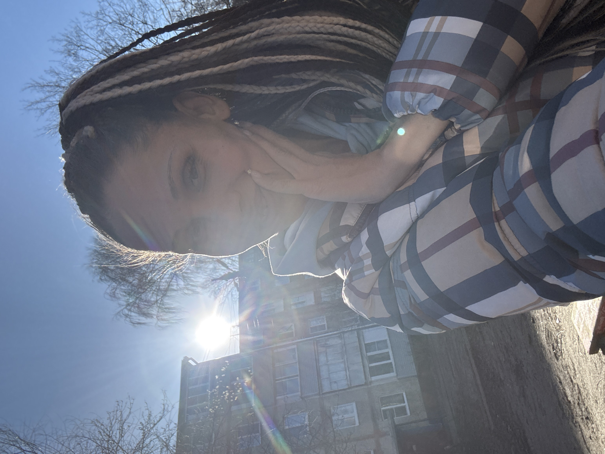 A brightly illuminated outdoor portrait shows a person with multi-toned braided hair, looking towards the camera from a low angle. The person's face, from the nose up, is visible, partially obscured by intense backlighting from the sun. Their right hand is gently positioned near their cheek or chin, revealing a manicured nail. They are wearing a plaid long-sleeved top or jacket in shades of blue, beige, and reddish-brown, with gathered or elasticated cuffs.
The background features a clear blue sky, against which bare tree branches are silhouetted, suggesting a colder season like late winter or early spring. Below the trees, a multi-story urban building with numerous windows is visible, bathed in the strong sunlight. The sun, positioned high in the sky, casts significant lens flares and a bright halo effect around the subject's head. The ground surface is visible in the lower right. The overall atmosphere is serene and sunny. The image was captured in Rusanovo, Russia. No visible text is present.