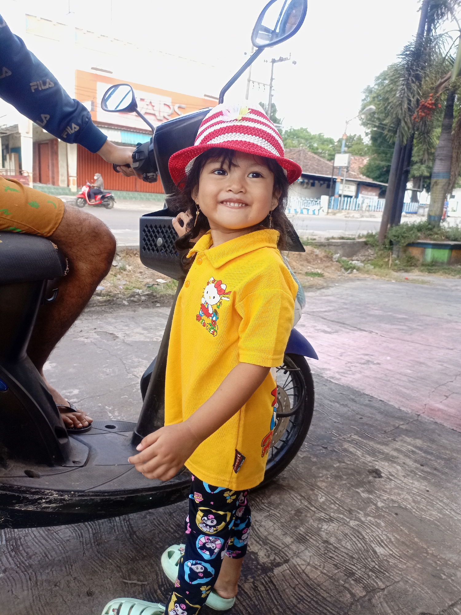 A young girl, appearing to be of Indonesian ethnicity, stands on a paved street in Juwana, Indonesia, next to a parked motorcycle. She is wearing a red and white striped straw hat, a yellow collared shirt with a Hello Kitty graphic, and patterned leggings featuring cartoon characters. Her left hand is slightly raised, and she is smiling. A person's arm, wearing a dark blue long-sleeved shirt with "FILA" logos, is visible on the left side of the frame, near the motorcycle's handlebars. The background shows a street with buildings, a red motorcycle with a rider in the distance, and palm trees. The ground is a concrete surface with some cracks and pinkish discoloration. The overall lighting suggests it is daytime.