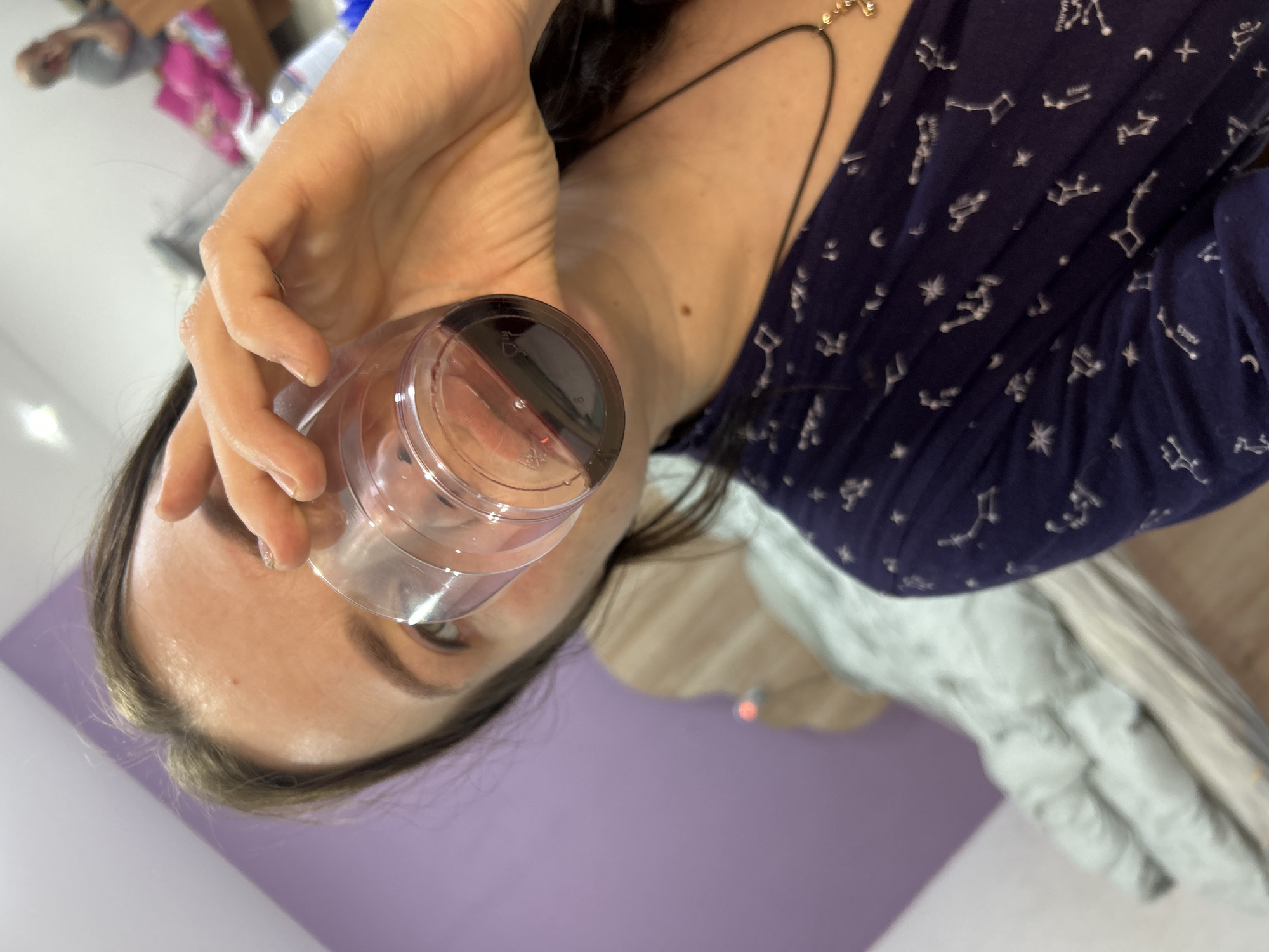 The image, captured in Rusalevka, Russia, shows a close-up, slightly angled view of a person, likely female, holding a clear plastic cup near their face. The person's head is tilted, with their forehead, well-defined eyebrows, and one eye partially visible above the cup. Their dark hair frames their face.
The individual is wearing a dark blue long-sleeved top. The fabric is patterned with numerous white designs representing constellations, stars, and crescent moons. A thin black cord necklace with a small golden, spherical charm or bead is visible around their neck. The person's hand firmly grips the clear plastic cup. The base of the cup displays typical recycling symbols and indistinct molding marks, commonly found on plastic containers. The contents of the cup are not discernible, but it appears to be empty or contains a clear substance.
The background is partially blurred but reveals an indoor setting. A section of a light purple wall is prominent on the left side. To the right and further back, a soft, light-colored fabric, possibly a blanket or garment, is draped over an unseen object. The upper background is very blurry, hinting at other indistinct objects or reflections, suggesting a domestic environment. The lighting is bright, indicative of a well-lit room or daytime. No specific emotions are clearly conveyed due to the angle and the cup obscuring much of the face.