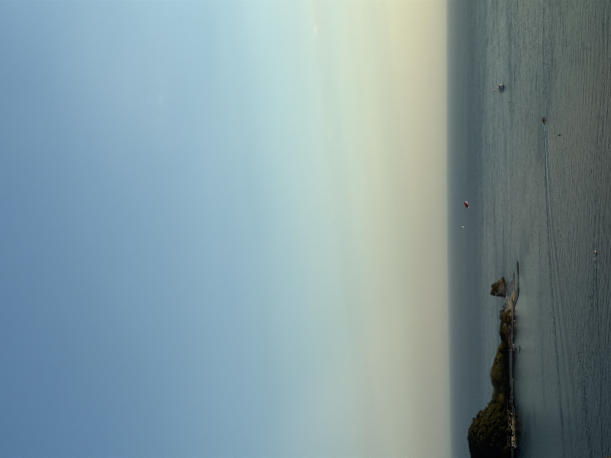 The image captures a serene coastal scene, likely in or near Boreti, Montenegro. The upper portion of the frame is dominated by a clear, gradient sky, transitioning from a light blue at the top to a pale yellow near the horizon, suggesting either sunrise or sunset.
Below the sky, the vast expanse of the Adriatic Sea stretches across the midground and foreground. The water appears calm with gentle ripples, reflecting the soft light of the sky. In the distance, a small, rocky island with lush green vegetation rises from the sea, connected to a more developed landmass by a narrow jetty or causeway. Several small boats are docked or anchored near this landmass.
Further out on the water, several objects are visible. A white boat is cruising on the left side of the frame, leaving a faint wake. In the mid-right, a parasailor with a red canopy is in the air, pulled by an unseen boat. Towards the far right, a small white vessel, possibly a yacht, is also navigating the water. The overall impression is one of peaceful leisure and natural beauty.