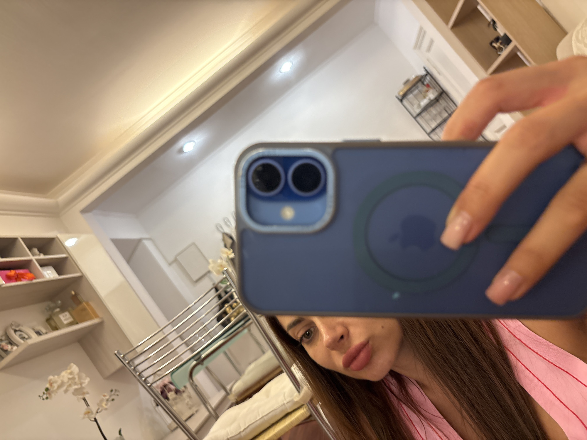 A woman takes a selfie with her phone in what appears to be a well-lit room with shelves and decorative items. The phone case is blue and has an Apple logo. She is wearing a pink top. The background shows a ceiling with recessed lighting and some shelves containing various objects.