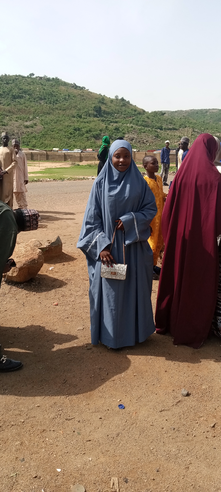 The image shows a group of people standing in an outdoor setting in Naraguta Hausawa, Nigeria. A woman in a blue hijab and long dress stands in the center, holding a small silver purse. To her right, a woman is mostly obscured, wearing a maroon head covering and dress. A child in a yellow robe stands behind the woman in blue. Several men in traditional clothing stand in the background to the left. A hill covered with vegetation is visible in the background. The ground is a mix of dirt and gravel. Other people are partially visible scattered around the scene. A low wall is visible behind them.