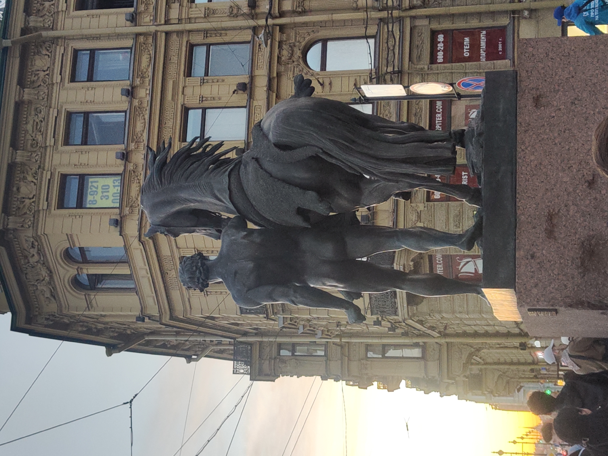 Here's a matter-of-fact description of the image:
The image depicts a statue in Saint Petersburg, Russia, specifically the "Taming of the Horse" sculptures on Anichkov Bridge. The statue features a nude male figure struggling to control a rearing horse. The statue is elevated on a stone base.
The backdrop shows a section of a building with multiple windows and ornate architectural details. Commercial signage is visible, with text including phone numbers ("600-20-80," "8-921 310 00-13") and words like "ОТЕЛИ" (hotels), "АПАРТАМЕНТЫ" (apartments), and "2PITER.COM."
In the lower right corner of the image are pedestrians. The lighting suggests late afternoon or early evening, with the sun low on the horizon. Power lines are also visible in the sky.