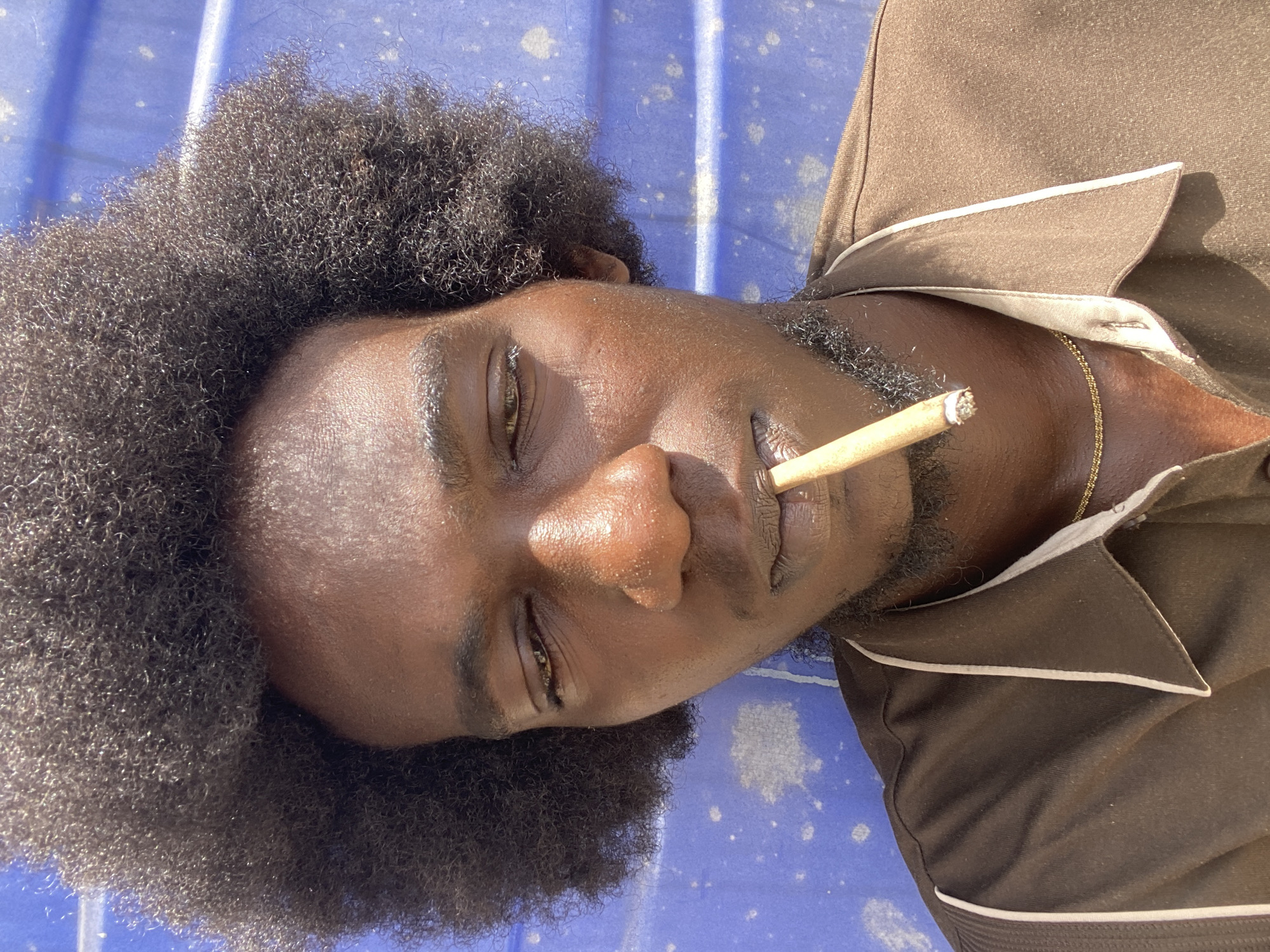 A man with dark skin and an afro is lying on his side on a blue surface. He is wearing a brown shirt with white trim and a gold chain. He has a cigarette in his mouth. The photograph was taken outdoors in Lagos, Nigeria.