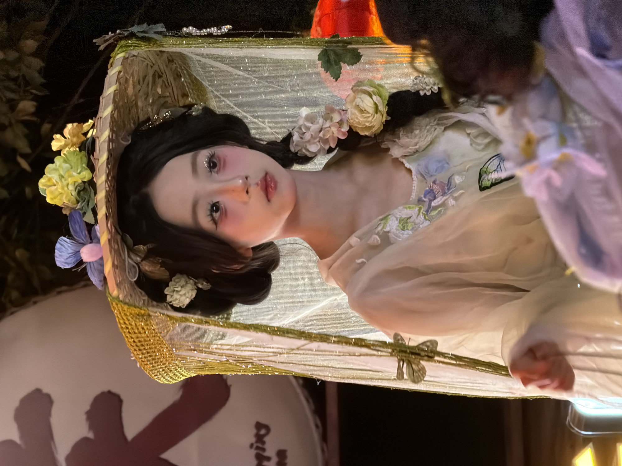 A young East Asian female wearing a traditional Chinese-inspired costume and an ornate hat adorned with flowers stands in what appears to be an outdoor setting at night. She has dark hair styled in a bun with a braid decorated with flowers. Her makeup features bold red blush and sparkling eyeshadow. The costume is light-colored and decorated with floral embroidery and a butterfly motif. She is partially obscured by sheer, shimmering fabric draped from her hat. In the background, a large, blurry red lantern and what appears to be Chinese calligraphy are visible. The scene is dimly lit, with some yellow and red light sources. The location is described as being in the Yuzhong District of China.