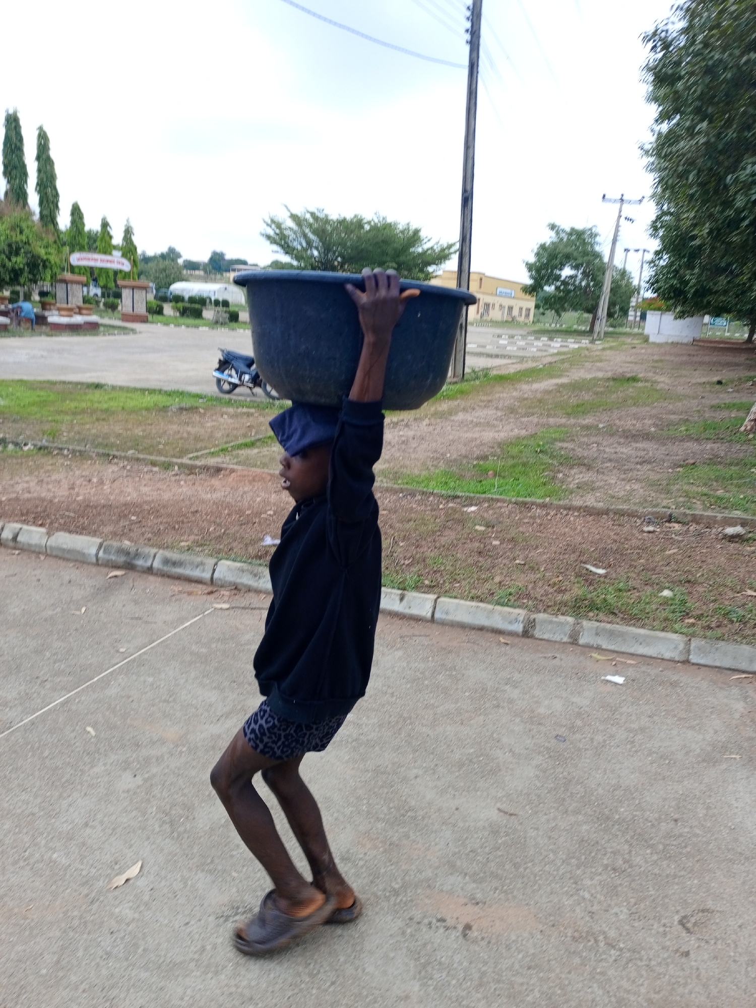 A person is carrying a large, dark-colored tub on their head. The person appears to be a child or young adult. The person is wearing a dark-colored top, patterned shorts, a dark cap, and sandals. They are walking on a paved road. The background shows a grassy area with trees, buildings, and utility poles. There is a parked motorcycle visible in the distance. The location appears to be in Keffi, Nigeria.