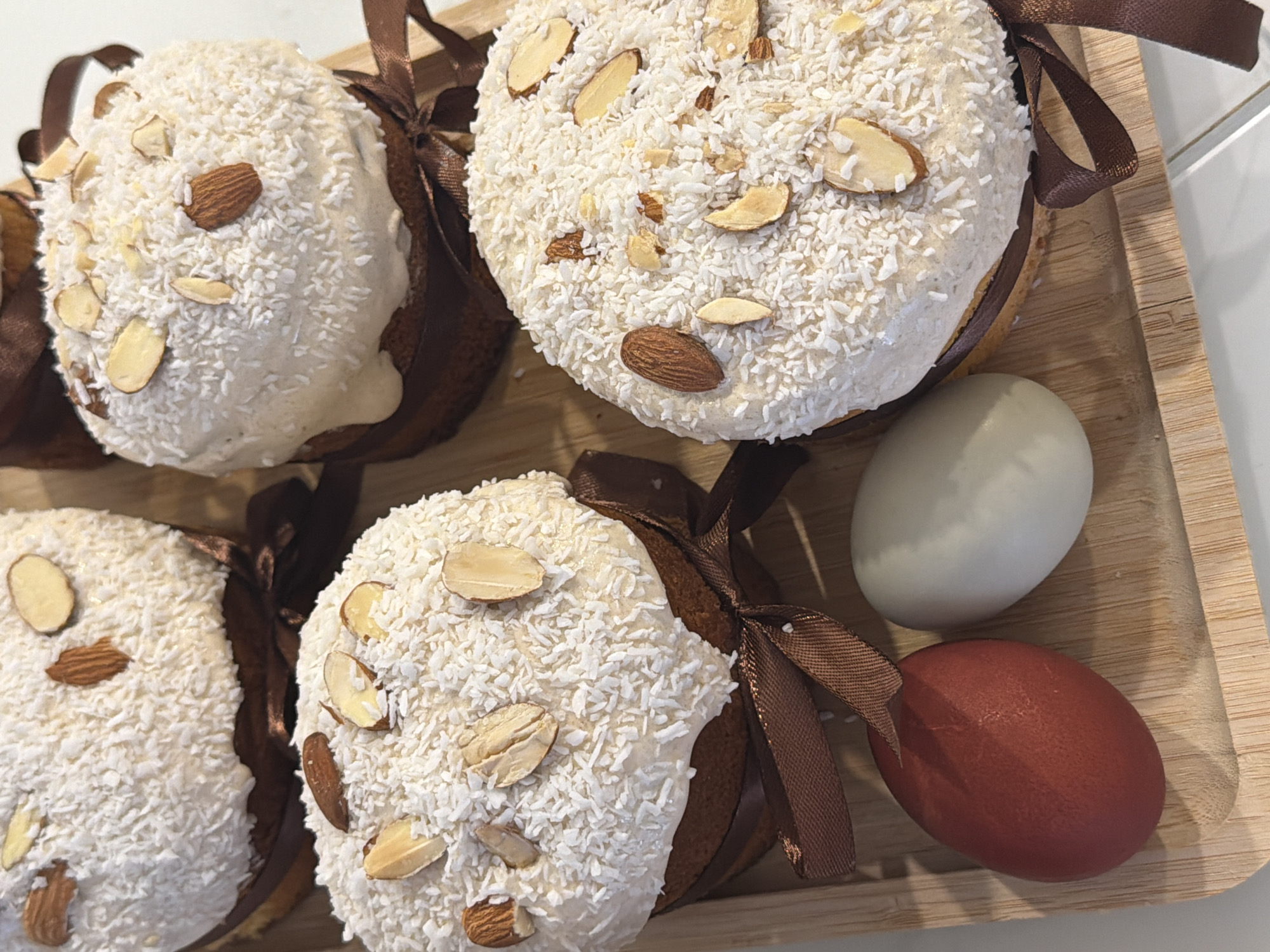 The image shows a wooden tray containing four Easter cakes and two Easter eggs. The cakes are round, covered with white frosting and shredded coconut, and decorated with sliced almonds. Each cake is tied with a brown ribbon. One egg is white, and the other is reddish-brown. The background appears to be a kitchen countertop or table. The location in Khimki, Russia is not apparent from the visual data.