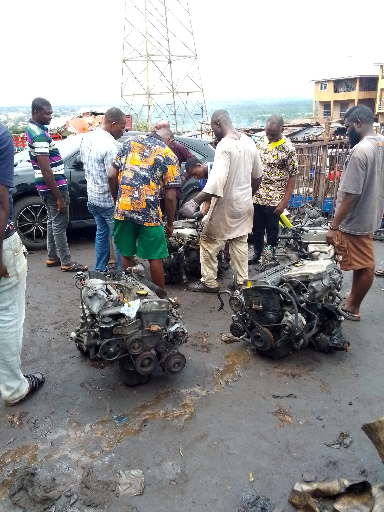 The image depicts a group of approximately eight men in an auto parts market in Onitsha, Nigeria. The ground is stained with oil and appears muddy. Two car engines are prominently displayed in the foreground. The men are dressed in a variety of casual clothing. A black car is parked nearby, with a utility tower visible in the background. Buildings and vegetation are visible in the distance. Some of the men appear to be inspecting the engines, possibly discussing their condition or value.