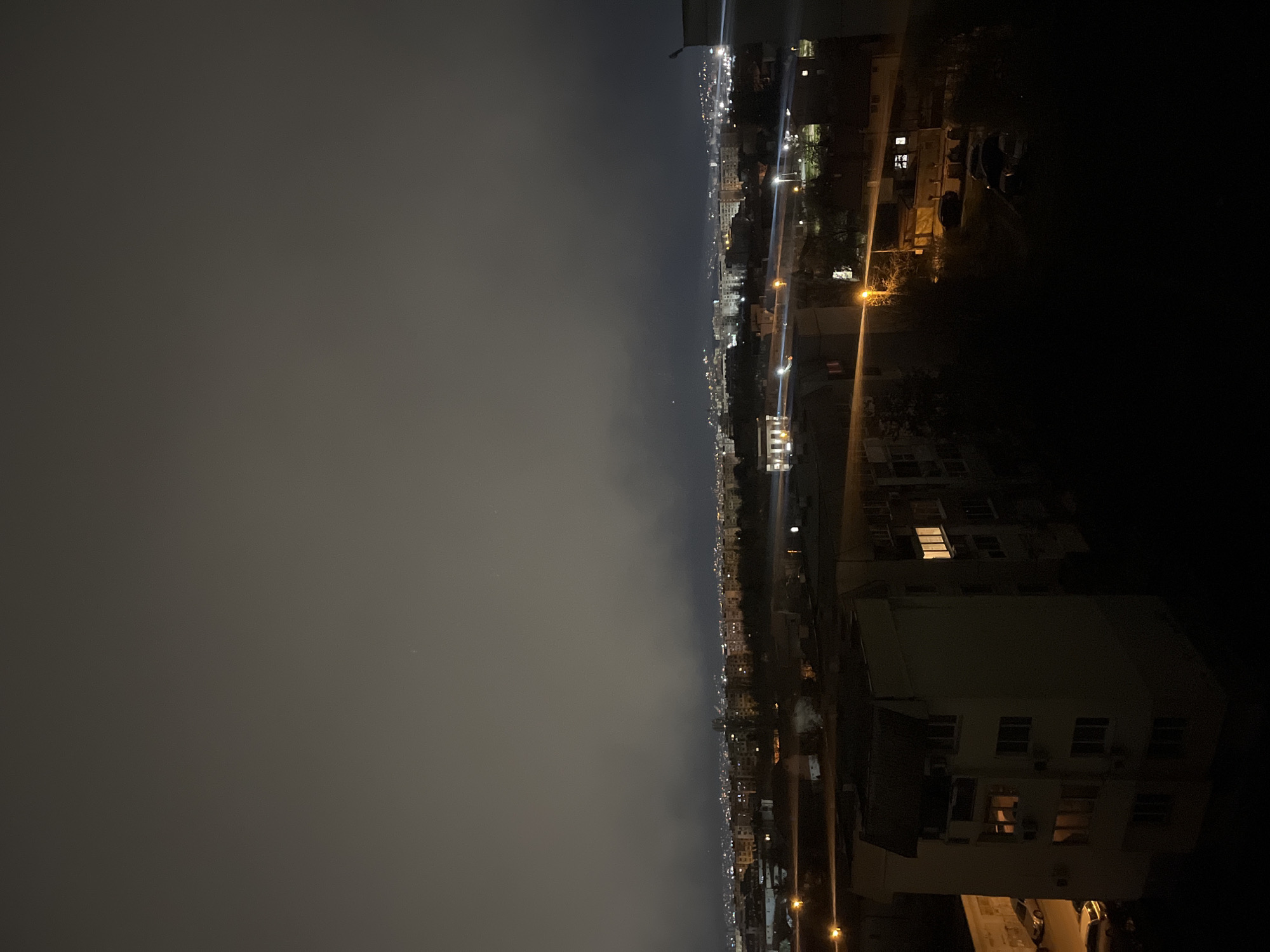 The image shows a nighttime cityscape, likely taken from a high vantage point in Sofia, Bulgaria. The right side of the image is dominated by buildings close to the camera, including a large multi-story apartment building with several lit windows. Streetlights are visible, illuminating the surrounding areas. The left side of the image depicts a more distant view of the city shrouded in a layer of fog or low clouds. City lights glimmer through the haze, suggesting a dense urban environment. The sky is dark and overcast.
