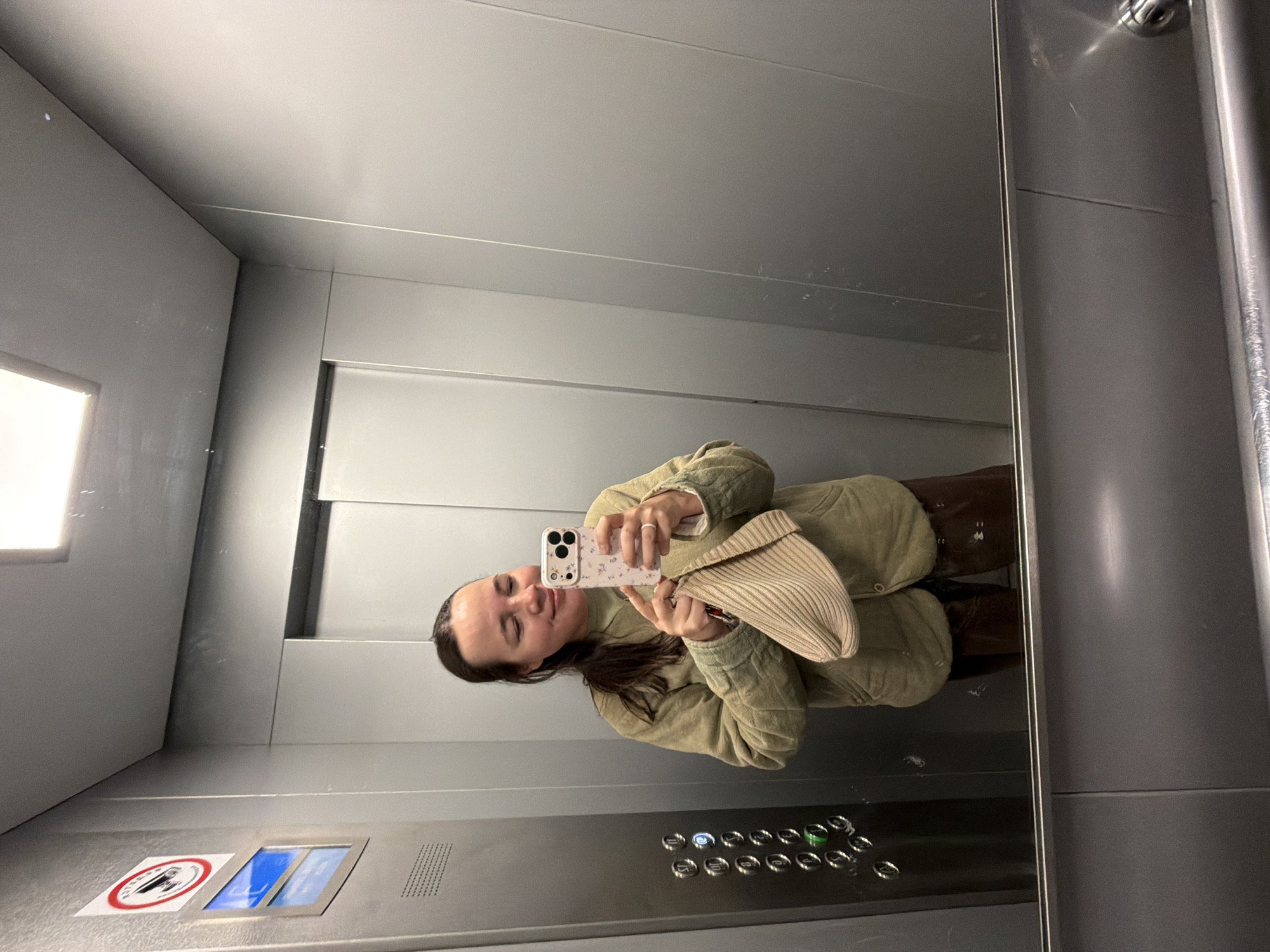 A woman is captured in a mirror selfie inside an elevator, holding her smartphone to take the picture. She is wearing a light olive green quilted jacket and holds a ribbed, light-colored clutch bag in her left hand. Her dark hair is pulled back, and she has a relaxed smile with slightly closed eyes. A ring is visible on her right hand.
The elevator interior is modern, characterized by metallic gray walls. A large mirror on the right reflects the woman and the opposite side of the elevator. The left wall features a square light fixture and a control panel towards the bottom. This panel is also metallic gray and includes numerous round buttons for floor selection, ranging from -1, 0, and 1 up to 20. One button, likely corresponding to floor 1, is illuminated with a green light. Below a rectangular display on the control panel, a "no smoking" sign (a red circle with a diagonal line over a cigarette) is visible, next to another small display showing a blue image. The smartphone used for the selfie has a case adorned with a subtle floral pattern.
This scene is set within an elevator in Chaikovsky, Russia.
