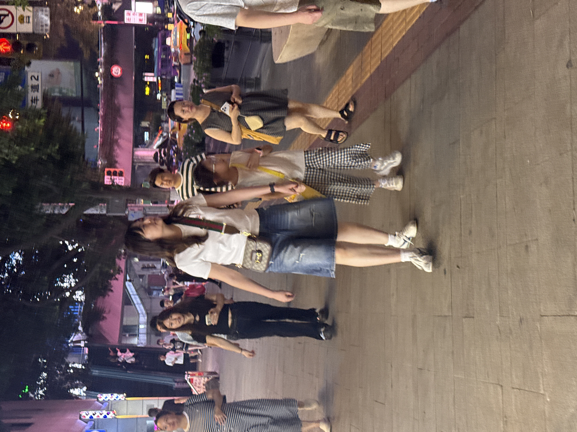 The image depicts a street scene in Yuzhong District, China, at night. Several people are walking on a paved sidewalk. In the foreground, a woman with long dark hair wears a white t-shirt and a denim skirt, with a brown and green patterned bag slung across her chest. To her left, a child in a white sleeveless top and black and white checkered pants walks, holding the woman's hand. To the right of the woman, another woman in a dark patterned dress and sandals is visible. In the background, other pedestrians, streetlights, building facades with illuminated signs, and passing vehicles are discernible. The environment suggests a bustling urban area with commercial activity.