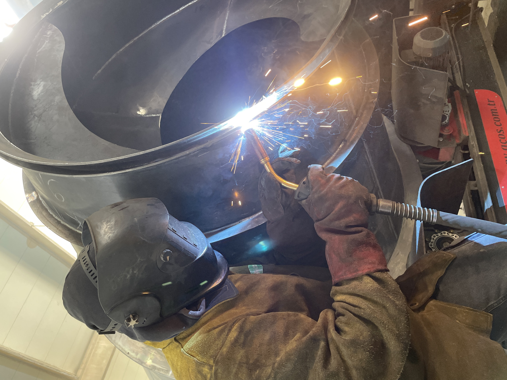 A person wearing a welding mask, brown gloves, and a brown jacket is welding a large, dark-colored metal structure with a curved, spiral shape. Bright sparks emanate from the welding torch, illuminating the surrounding area. To the right of the metal structure, a dark motor and a red and white sign bearing text that ends in ".com.tr" are visible. The scene is indoors, likely in a workshop or industrial setting, with a bright ceiling visible above. The location is Čeluga, Montenegro.