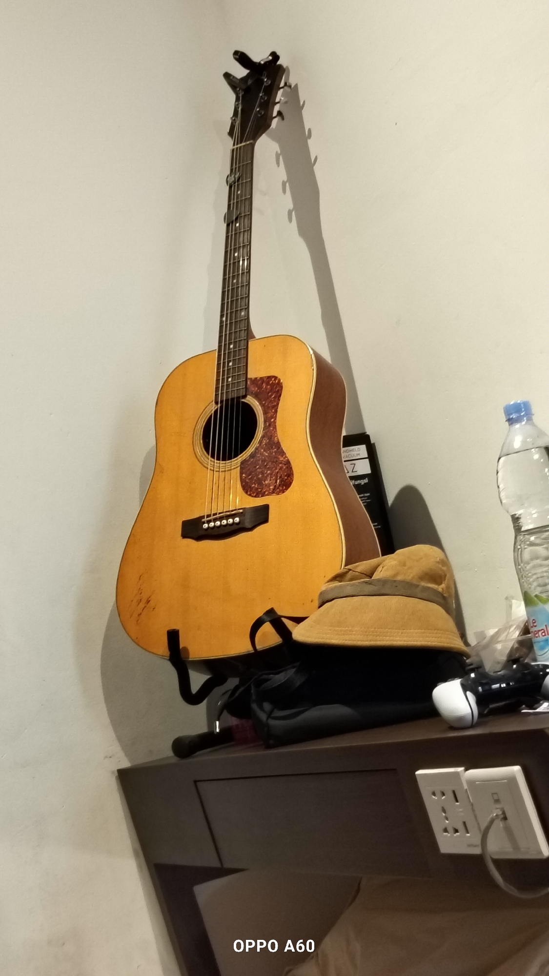 An acoustic guitar is displayed on a wall mount. Below the guitar, on a dark brown surface, are a brown bucket hat, a black bag, a white and black video game controller, and a clear plastic bottle of water. The wall is painted a light, neutral color. Electrical outlets are visible on the right side of the frame, with a cable plugged into one of them. The scene appears to be indoors, likely in a residential or hotel room. The image is tagged with "OPPO A60" which suggests the device used to capture the photograph. The overall impression is of a personal space with musical and electronic equipment. The location is specified as North Kuta, Indonesia.