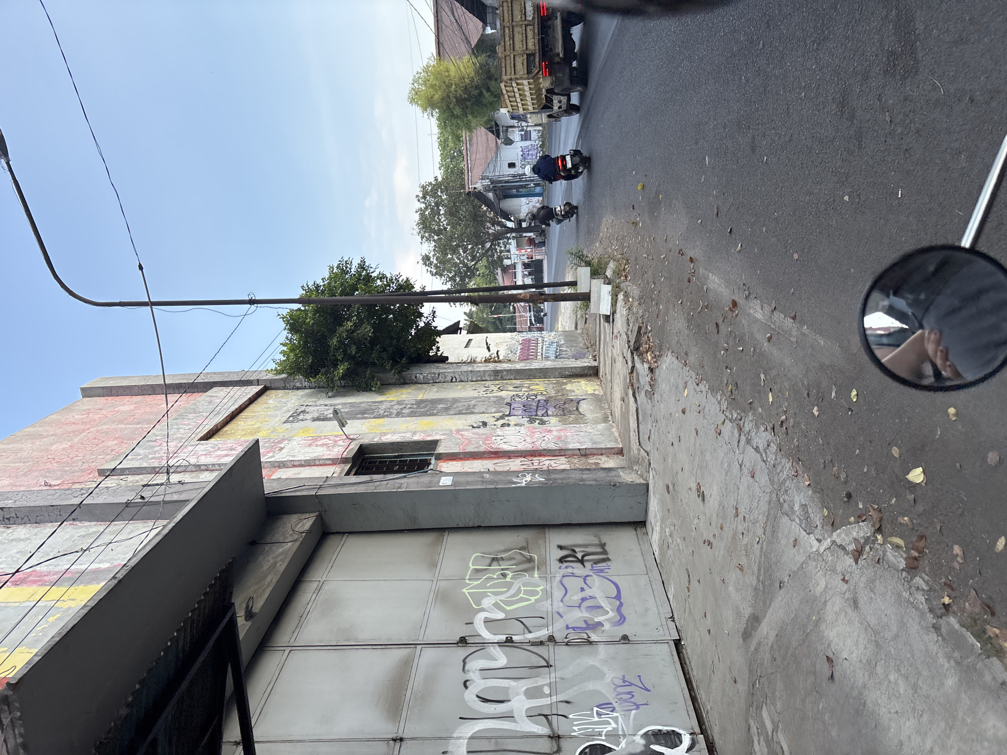 The image shows a street scene in Cirebon, Indonesia. In the foreground, a gray metal gate, covered in graffiti, is visible on the left. The building behind the gate is weathered and also shows signs of graffiti. A sidewalk runs alongside the building, littered with fallen leaves. The street itself is asphalt, with some leaves scattered on it. On the right side of the street, two motorcycles are being ridden, with a truck in front of them. Buildings with tiled roofs line the street in the background. The sky is clear and blue. The photo appears to be taken from the perspective of someone riding a motorcycle, as a rearview mirror is visible in the lower right corner.