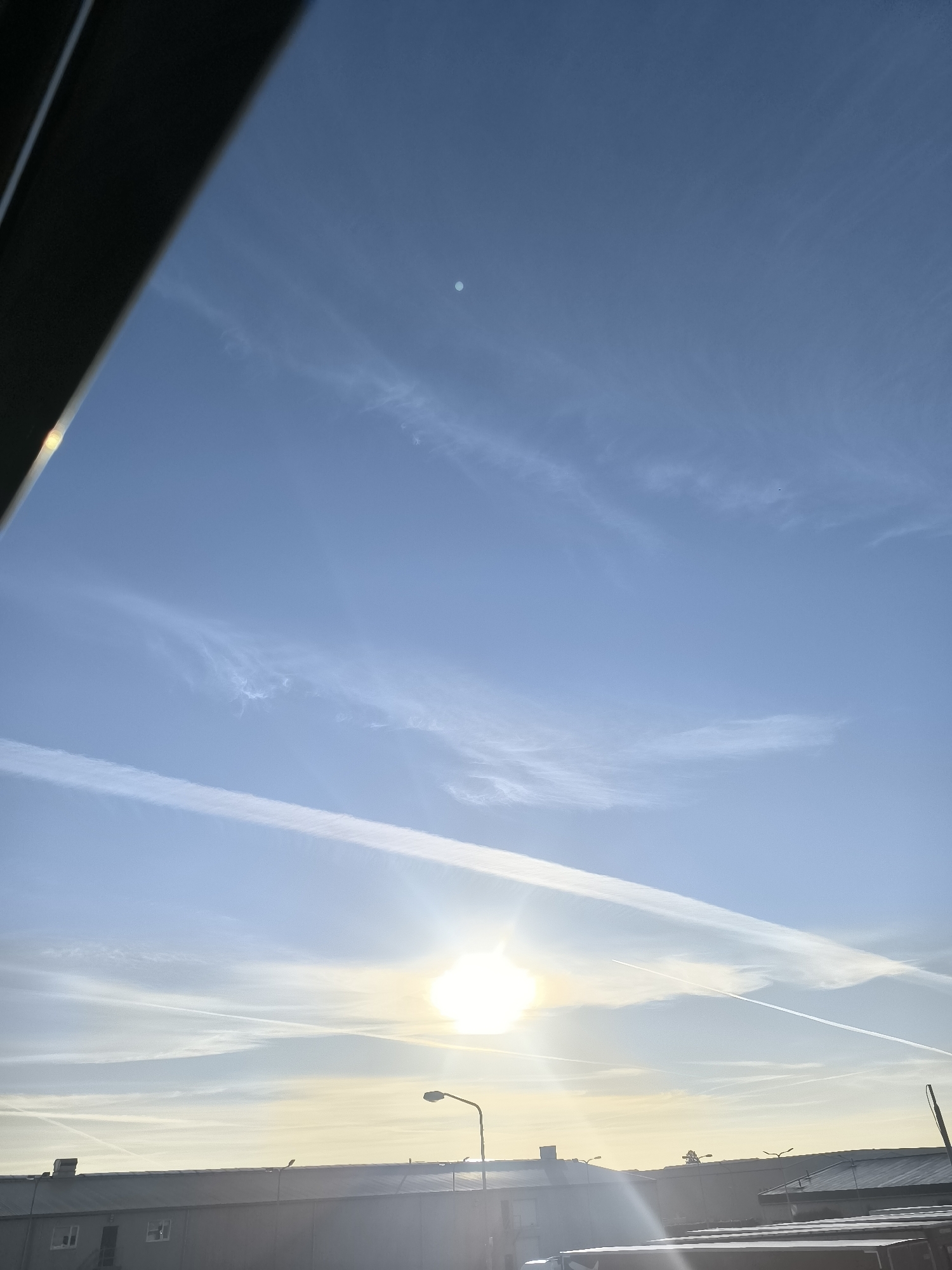 This image captures a bright, sunny sky over Uithoorn, Netherlands. The sun is prominently featured, casting a bright glare. Several contrails from airplanes streak across the clear blue sky, some appearing wispy and others more defined. In the lower portion of the image, the rooftops of industrial buildings are visible, with a streetlamp and other structures suggesting an urban or semi-industrial environment. The overall impression is one of a clear day with ongoing air traffic above the Dutch landscape.