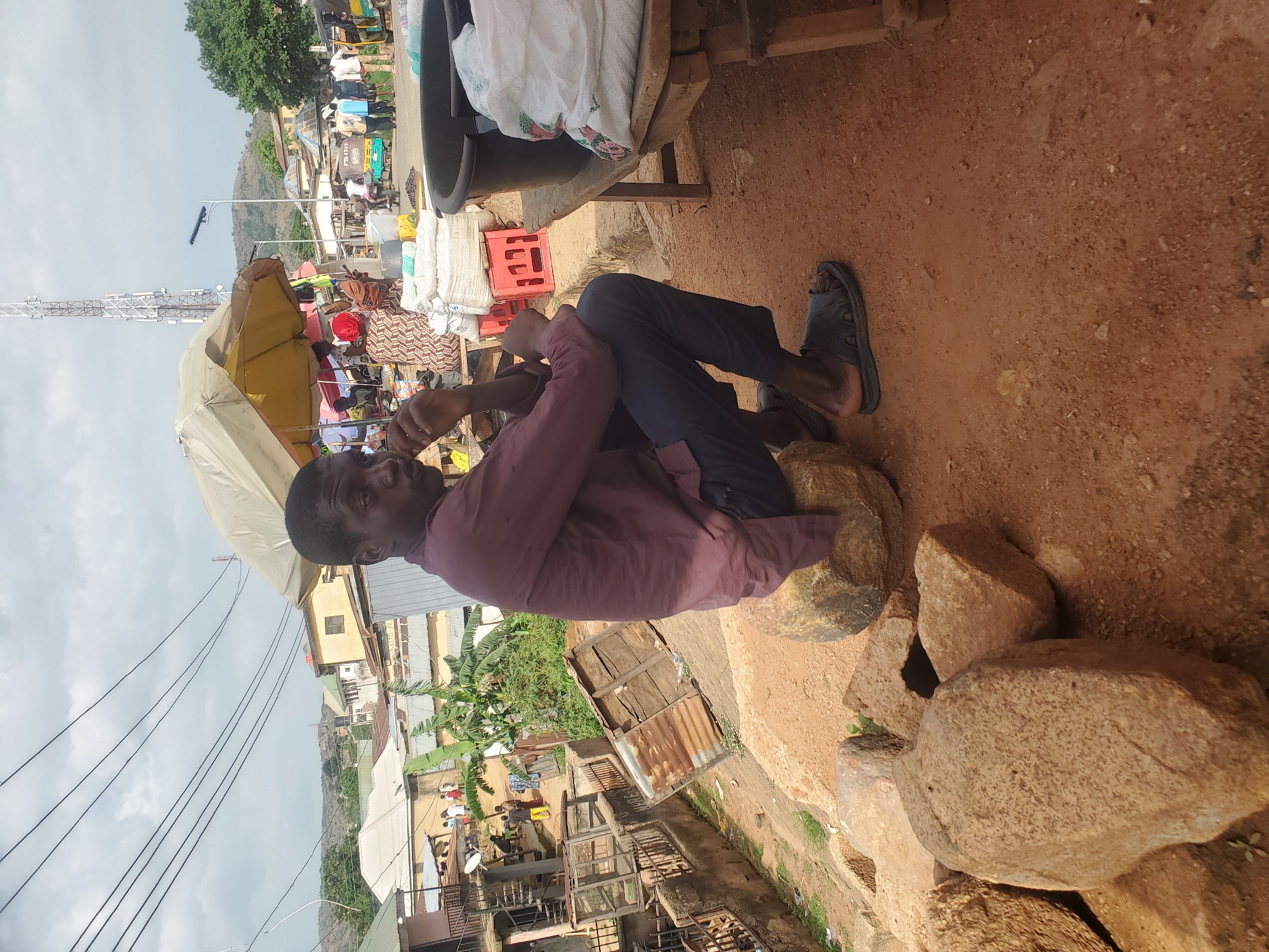 In the city of Gwong, Nigeria, a man sits on a large rock in an outdoor market. He is wearing a maroon long-sleeved shirt and dark pants, with black sandals on his feet. He is looking to his right, with his hand near his mouth.
Behind the man, a market scene unfolds. A yellow umbrella shades some goods, including white bags and red crates. A wooden bench holds more white bags and a large black basin. In the background, there are various buildings, some with corrugated metal roofs, and lush green trees. A tall telecommunications tower stands prominently against a cloudy sky. Several people are visible in the distance, some walking and others interacting. The ground is a reddish-brown dirt.