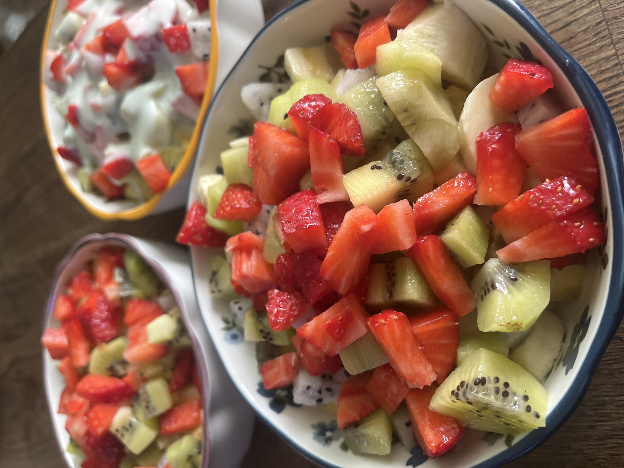 The image shows three bowls of fruit salad on a wooden surface. The largest bowl, centered and in focus, contains chunks of strawberries, kiwi, dragon fruit, and banana. A smaller bowl on the lower left contains a similar mix of strawberries and kiwi. The bowl in the upper left contains the same fruits coated in a white, creamy substance, likely yogurt. The scene is brightly lit. The location is unknown and cannot be determined from the image. The image does not include any people, actions, or interactions.