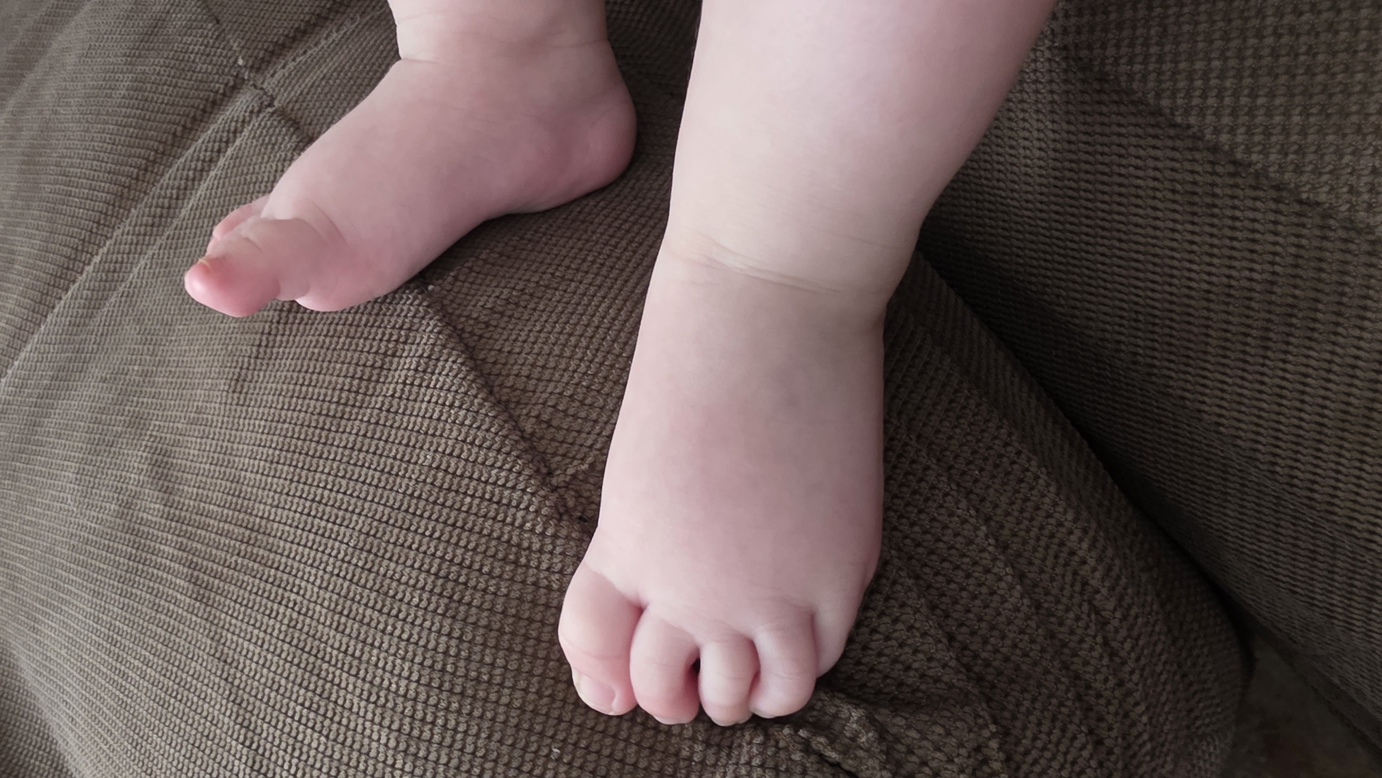 A close-up shows two baby feet resting on a textured, brown fabric surface, possibly a couch or similar furniture. The skin is pale pink. The location is not discernible from the image.