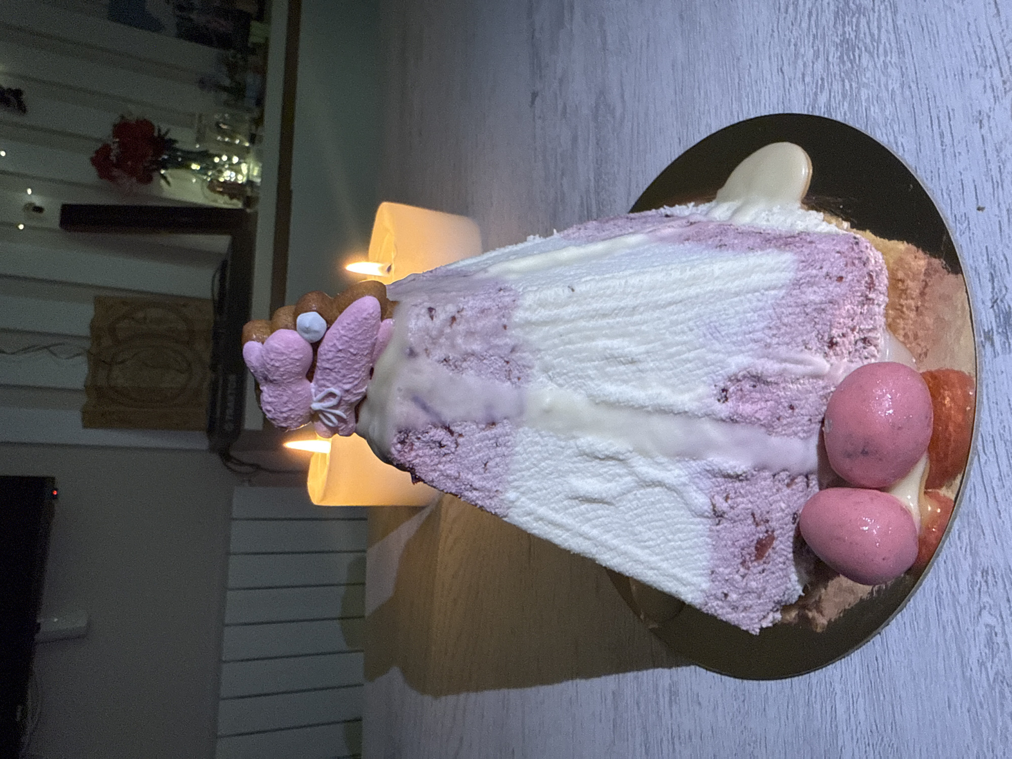 A photograph shows a large slice of cake on a gold plate. The cake has alternating layers of white and light purple sponge, with a white icing drizzle over the top. At the top edge of the cake are pink marshmallows and a gingerbread figure. The slice is also decorated with strawberries and pink icing balls. Two lit candles are positioned behind the cake. The background contains a shelf with a vase of red flowers and string lights. In the lower left corner, a television screen is visible. The floor is light brown and the wall is a light, bluish-grey. The photo was likely taken indoors, in a residential space in Batumi, Georgia.