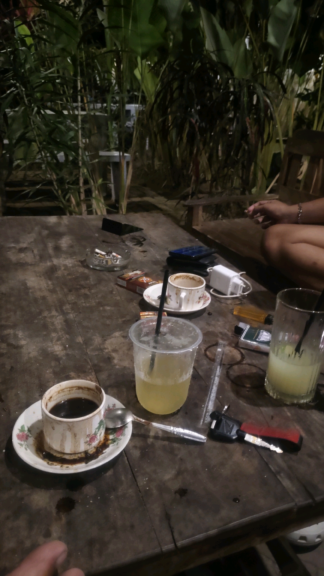 The image depicts a close-up, low-angle shot of a rustic wooden table at an outdoor cafe in Karangtalun, Indonesia, likely during the evening. The table is cluttered with various items, suggesting a casual gathering or a pause in activity.
On the table, there are two cups of beverages, one appearing to be dark coffee in a decorative ceramic cup with a saucer, and the other a clear, yellowish drink in a plastic cup with a straw. Nearby, there is an empty ceramic cup and saucer, also with remnants of a drink. Among the other items are a pack of cigarettes, an ashtray containing several cigarette butts, a lighter, a set of keys with a red and black fob, a mobile phone, and what appears to be a small white electronic device, possibly a charger.
In the background, lush green foliage creates a natural, almost jungle-like ambiance, with hints of artificial lighting suggesting a nighttime setting. A portion of a wooden chair and the legs of a person are visible on the right side of the frame, indicating the presence of at least one individual. The overall impression is one of a relaxed, unpretentious environment where people gather for drinks and conversation.