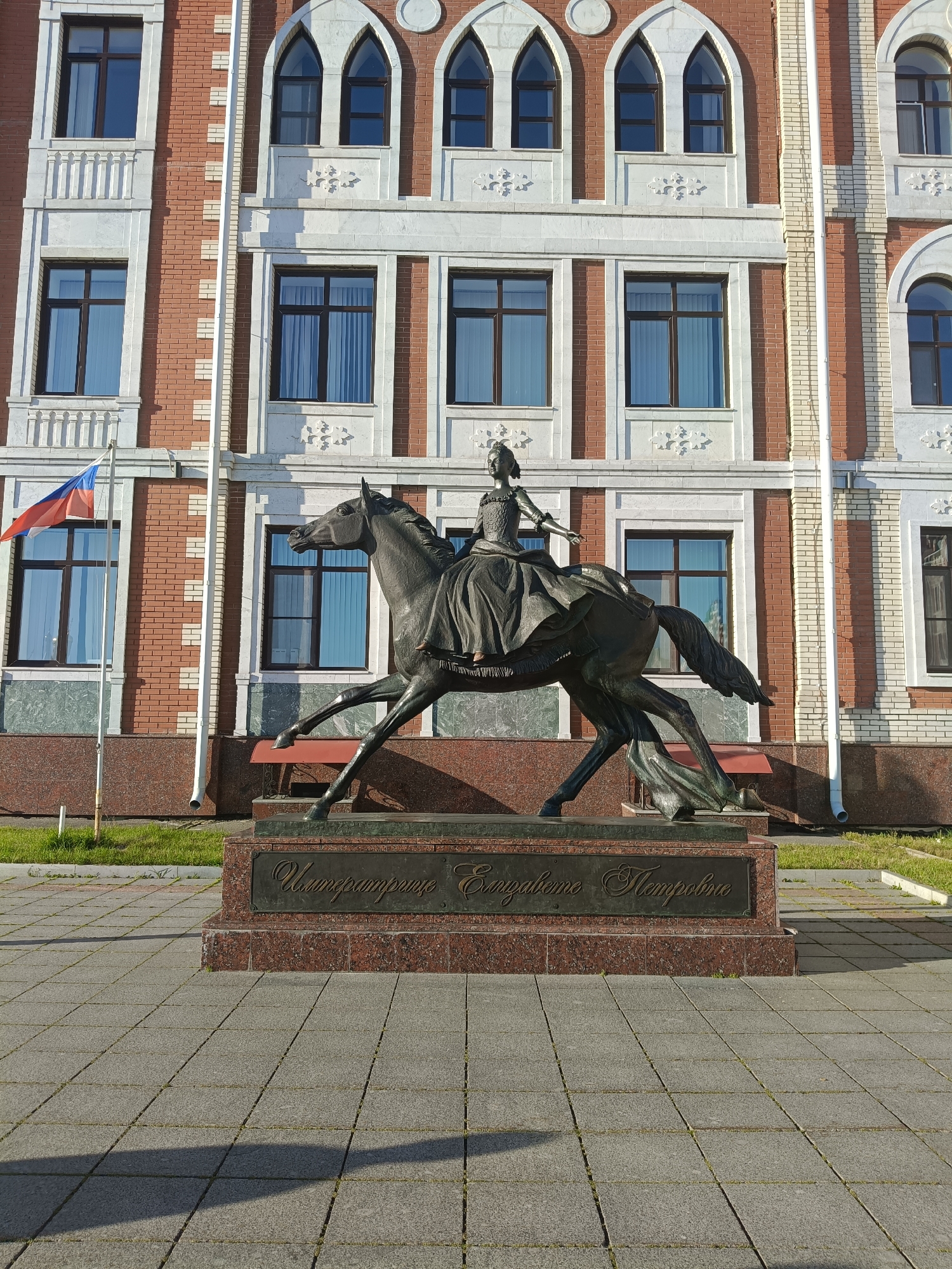 In the city of Yoshkar-Ola, Russia, a bronze equestrian statue of Empress Elizabeth stands in front of a red brick building with white architectural details and numerous windows. The statue depicts a woman in a flowing dress and crown, seated on a horse that appears to be in motion. The base of the statue features a plaque with an inscription in Russian. To the left of the statue, a flagpole flies the Russian flag. The area in front of the building is paved with square tiles, and a strip of green grass is visible along the base of the building. The scene is bathed in sunlight, casting shadows on the pavement.