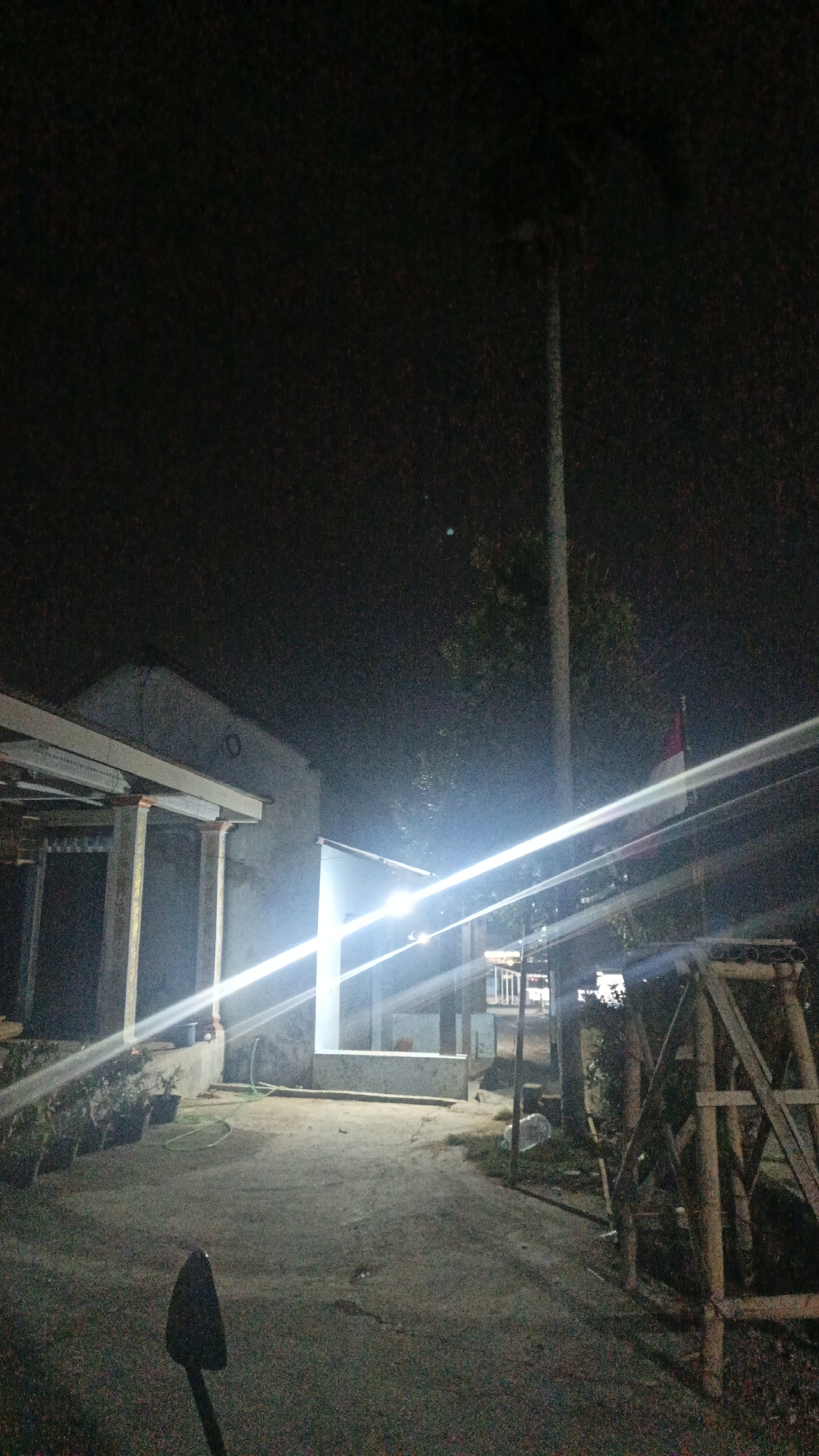 The image depicts an outdoor scene at night, possibly in Sambi, Indonesia, featuring a pathway leading towards a building. The building appears to be a simple structure with a covered porch or overhang. Bright artificial lights illuminate the area, creating strong glare and lens flares that streak across the image. To the right of the pathway, there is a wooden structure, possibly scaffolding or a support frame. A flag, predominantly red and white, is visible on a flagpole behind the wooden structure. The ground is a mix of concrete and dirt, with some vegetation and a plastic bottle scattered on the side. In the foreground, a dark, shovel-like object is positioned upright. The overall atmosphere is dimly lit, with the artificial lighting being the dominant source of illumination.