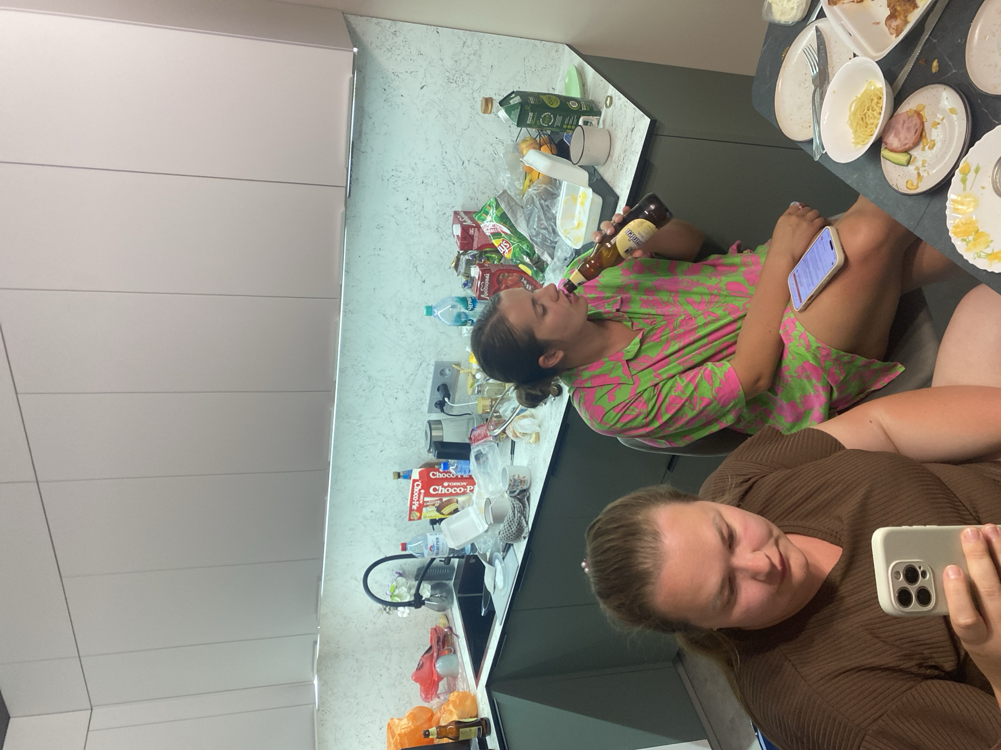 Two women are seated at a table in what appears to be a kitchen. The woman on the left is looking at her phone. The woman on the right is holding a bottle of beer to her lips. There are several plates with food remnants on the table, as well as various kitchen items and food packaging on the counter behind them. The kitchen features modern cabinetry and a speckled countertop. The scene suggests a casual gathering.