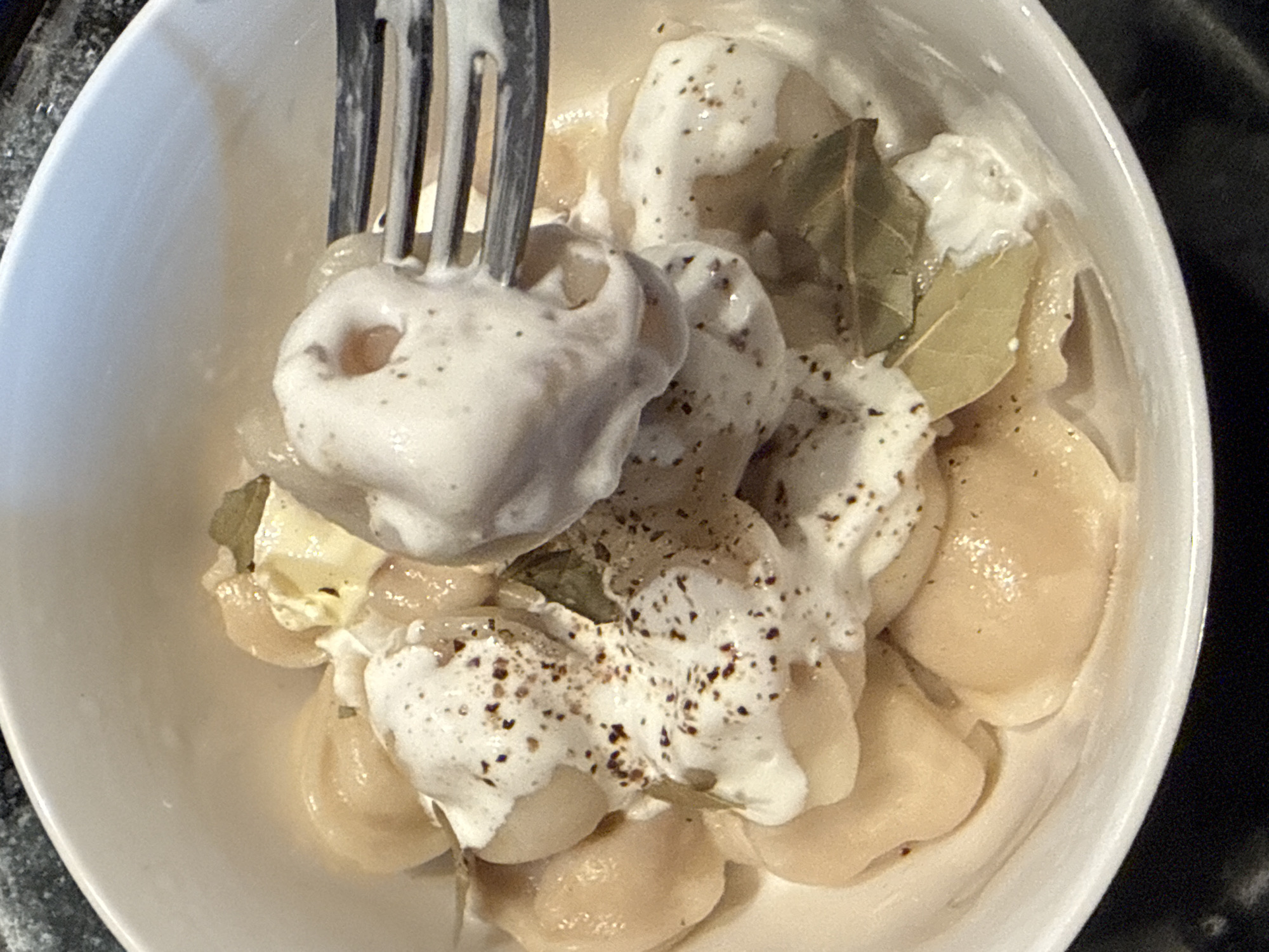 A close-up shot shows a white bowl filled with dumplings, likely pelmeni, in a creamy sauce, possibly sour cream or yogurt, and seasoned with black pepper. A fork is lifting a dumpling covered in the sauce. Bay leaves are visible among the dumplings. The setting is likely a home kitchen in Шолохово, Russia, given the context.